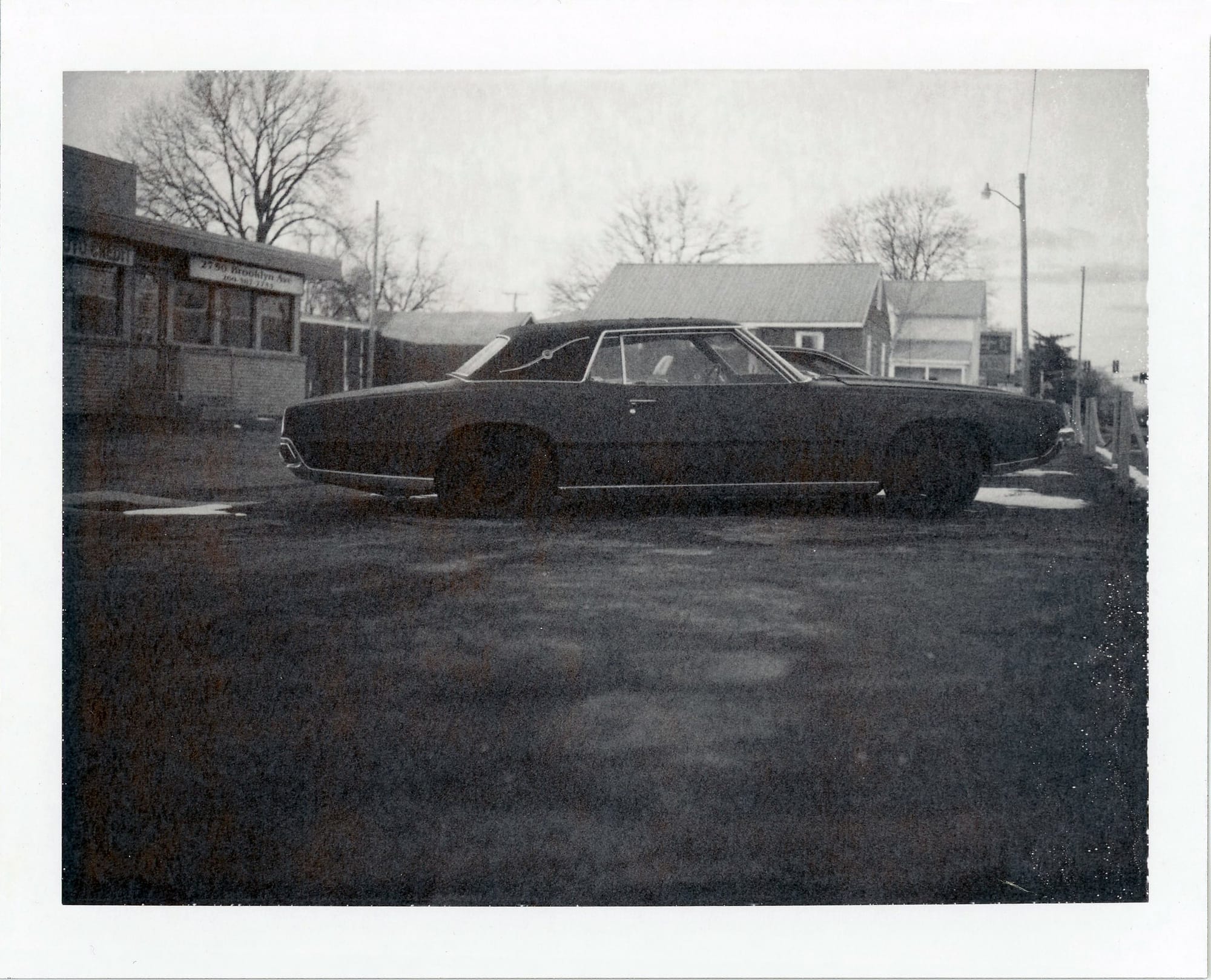 Dark muscle car with long hood parked in urban lot beside brick building on winter afternoon.