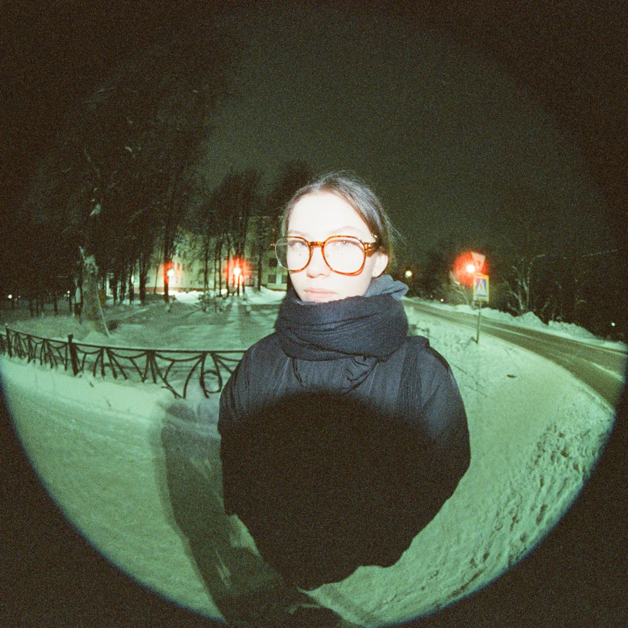 Young woman in glasses and winter outfit standing in a snowy park at night.