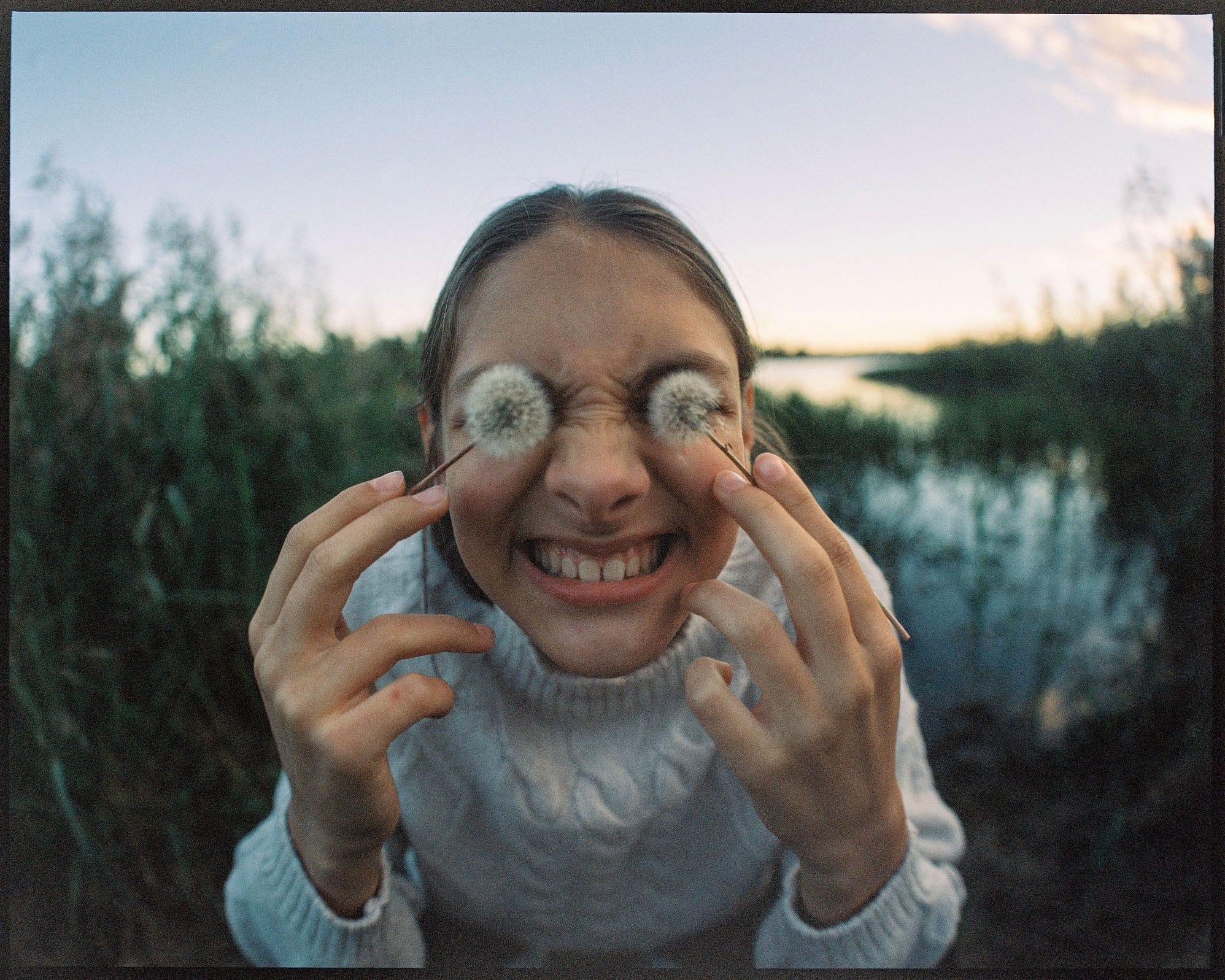Girl enjoying nature with dandelion fluff on her eyes, smiling in a serene outdoor setting.