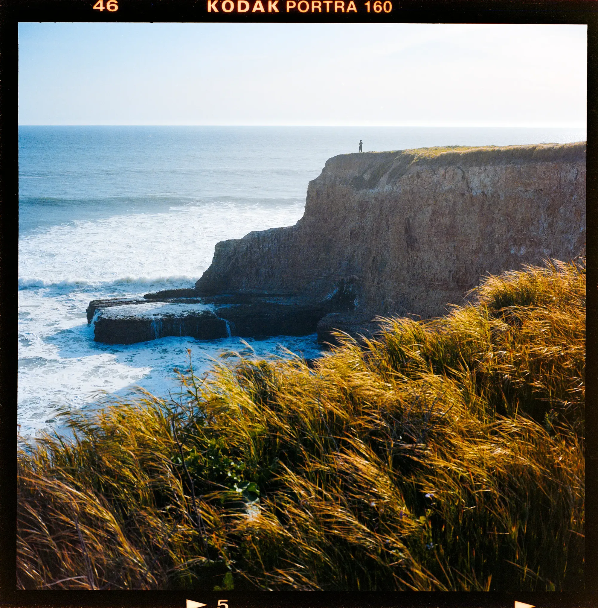 Person standing on coastal cliff overlooking ocean waves on Kodak Portra film.