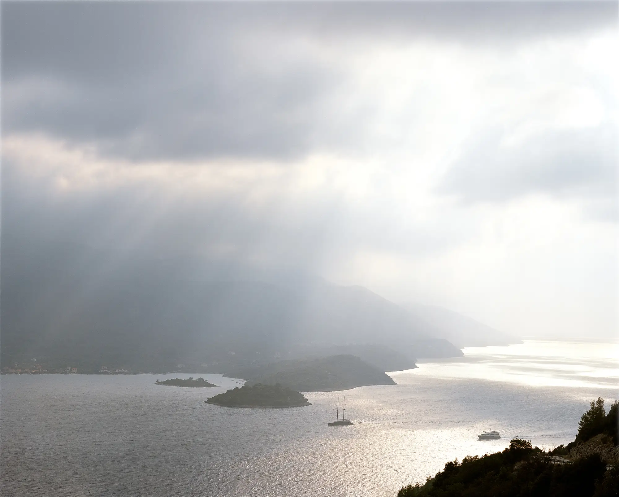Dramatic storm clouds over silver water with small islands and sailboat silhouetted against distant mountain peninsula.