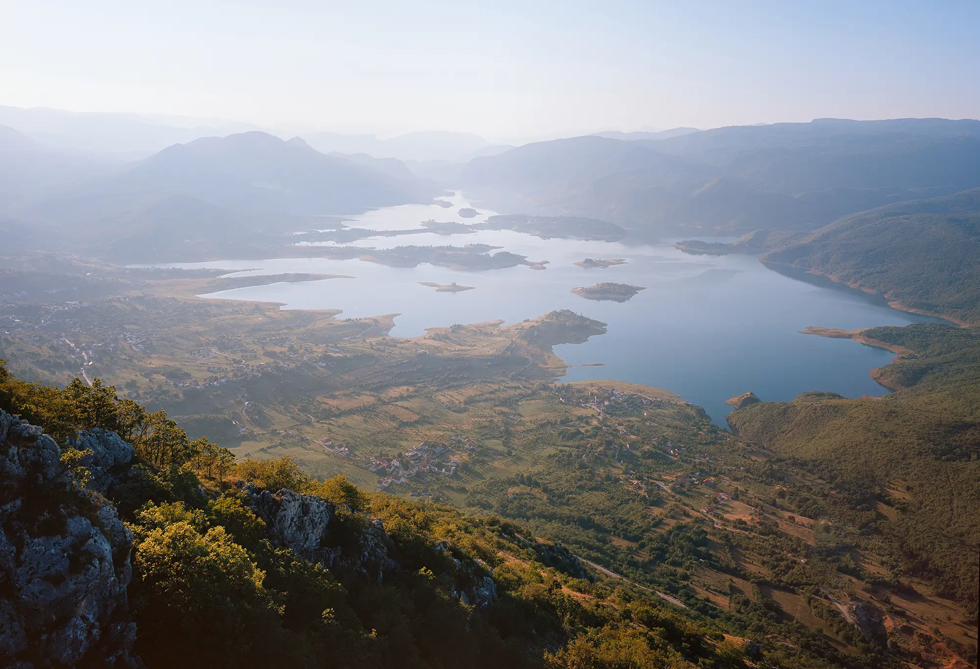 Aerial view of reservoir with multiple islands and scattered agricultural fields viewed from mountain summit under hazy light.