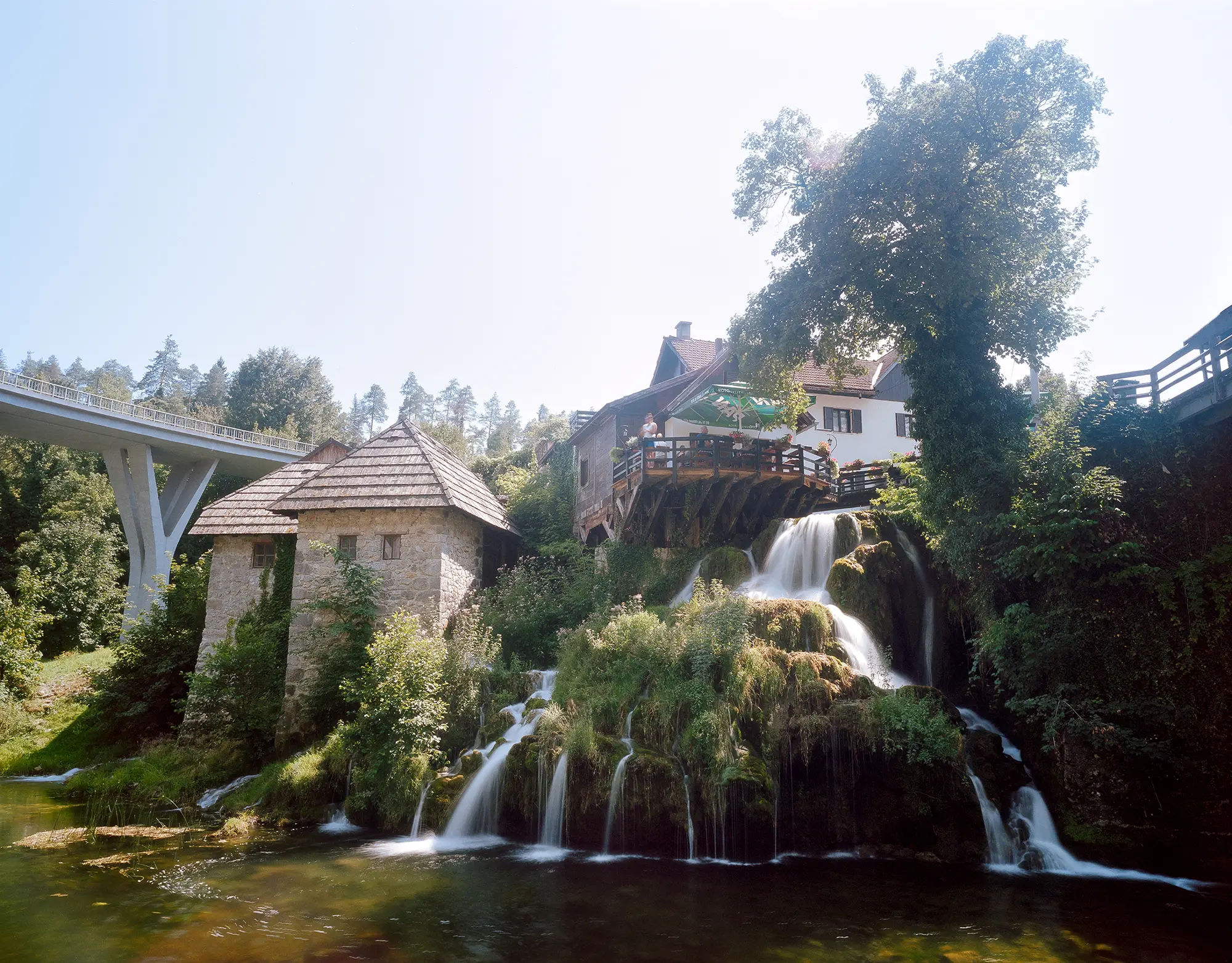 Historic stone mill buildings beside cascading waterfall with residential structures and white bridge under clear sky.