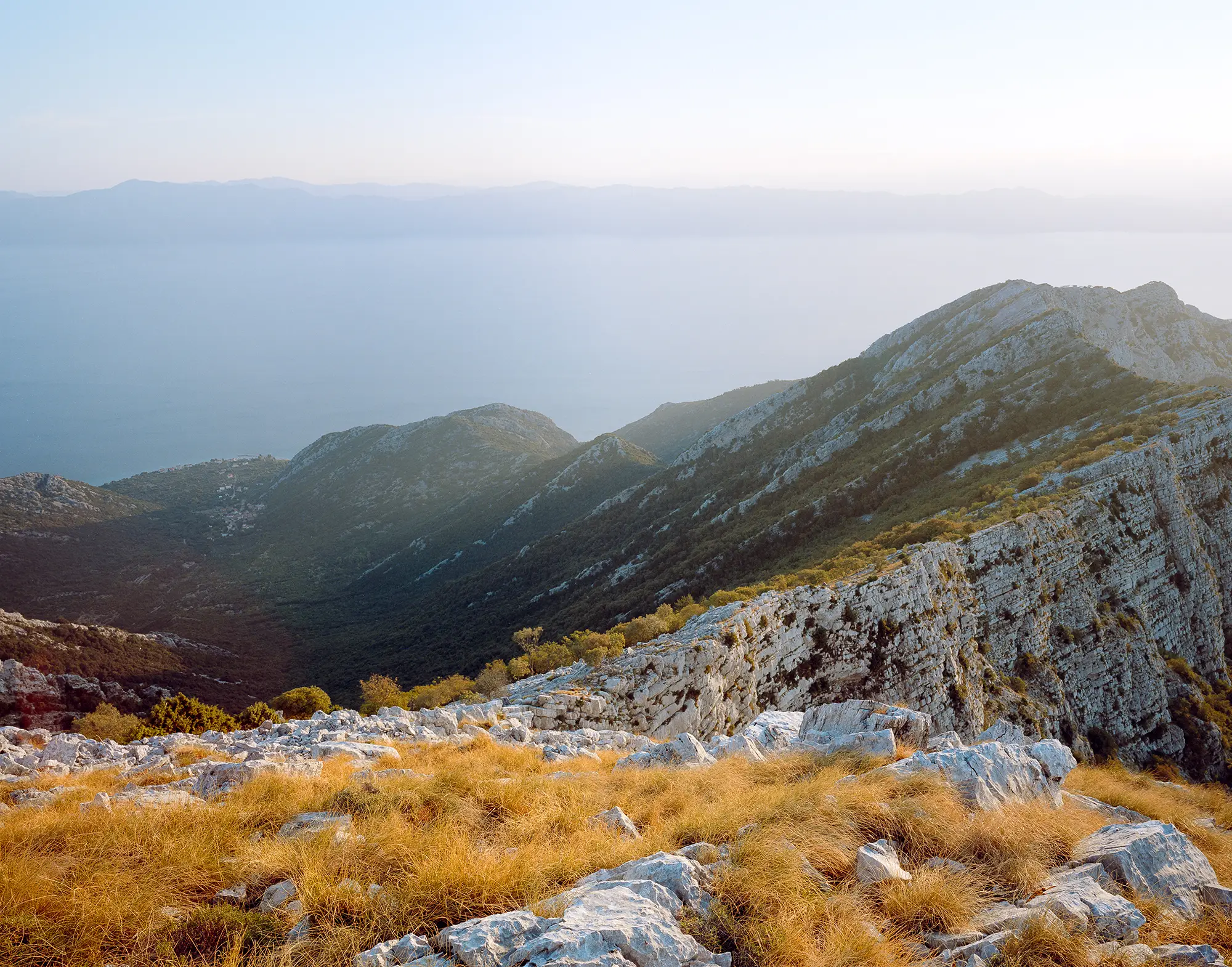 Rocky mountain ridge with golden dried grasses and white limestone outcrops descending toward misty valley at sunset.