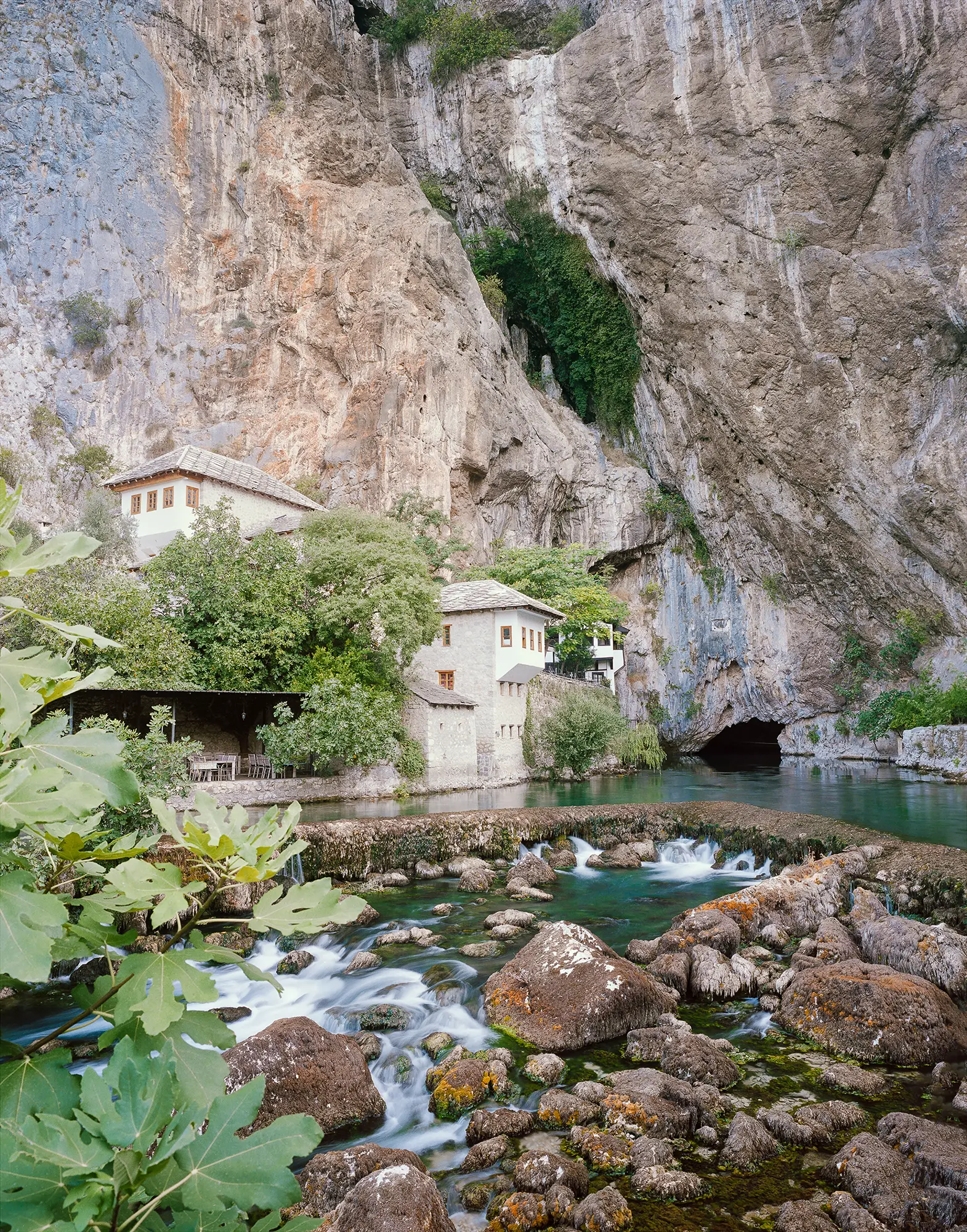 White Ottoman-style monastery buildings nestled under towering limestone cliff beside turquoise river with rushing stream.