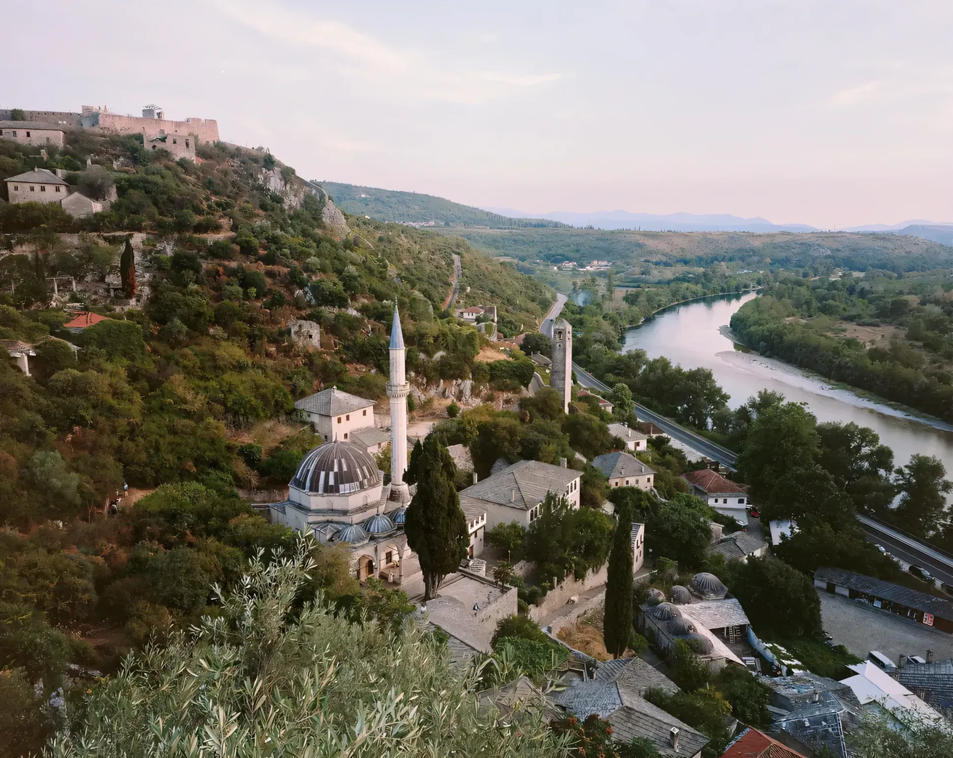 Hilltop fortress and mosque overlooking river valley at sunset with agricultural terraces and distant mountain ranges.