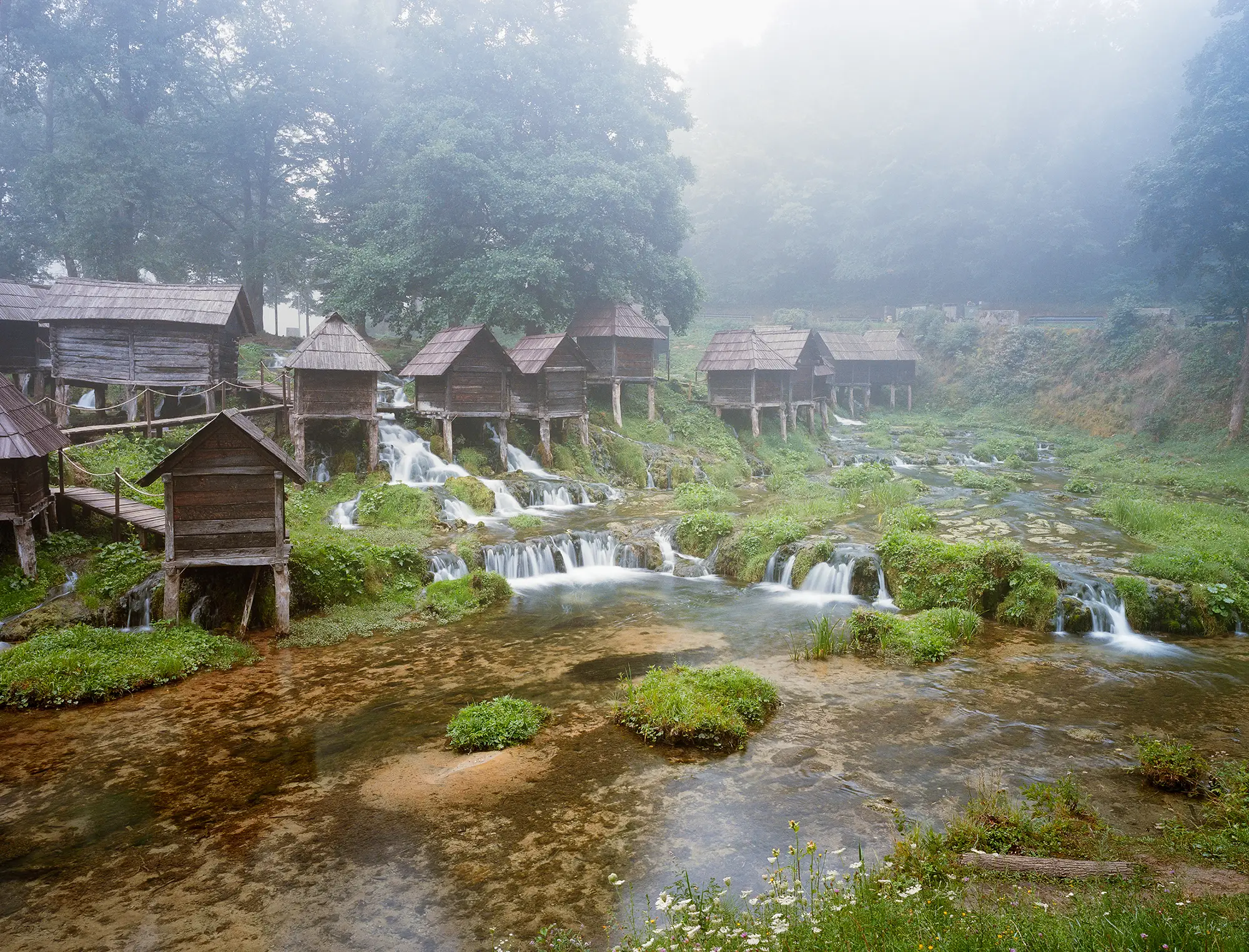 Traditional wooden watermills on stilts over cascading stream surrounded by lush vegetation in morning mist.