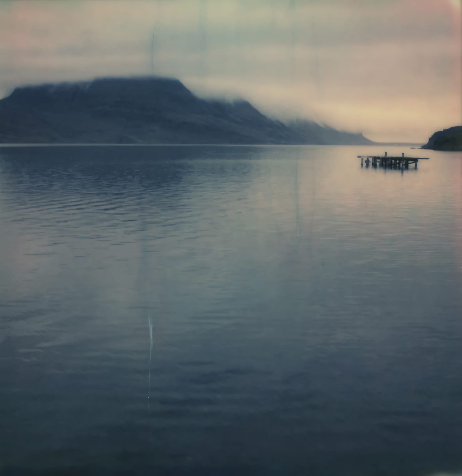 Wooden dock extending into calm fjord water with mountains in background on Polaroid.