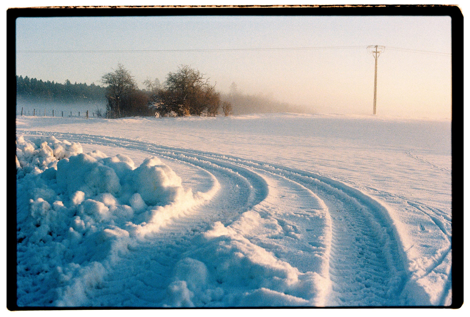 Curved tire tracks through deep snow leading toward bare trees and power line in winter field.