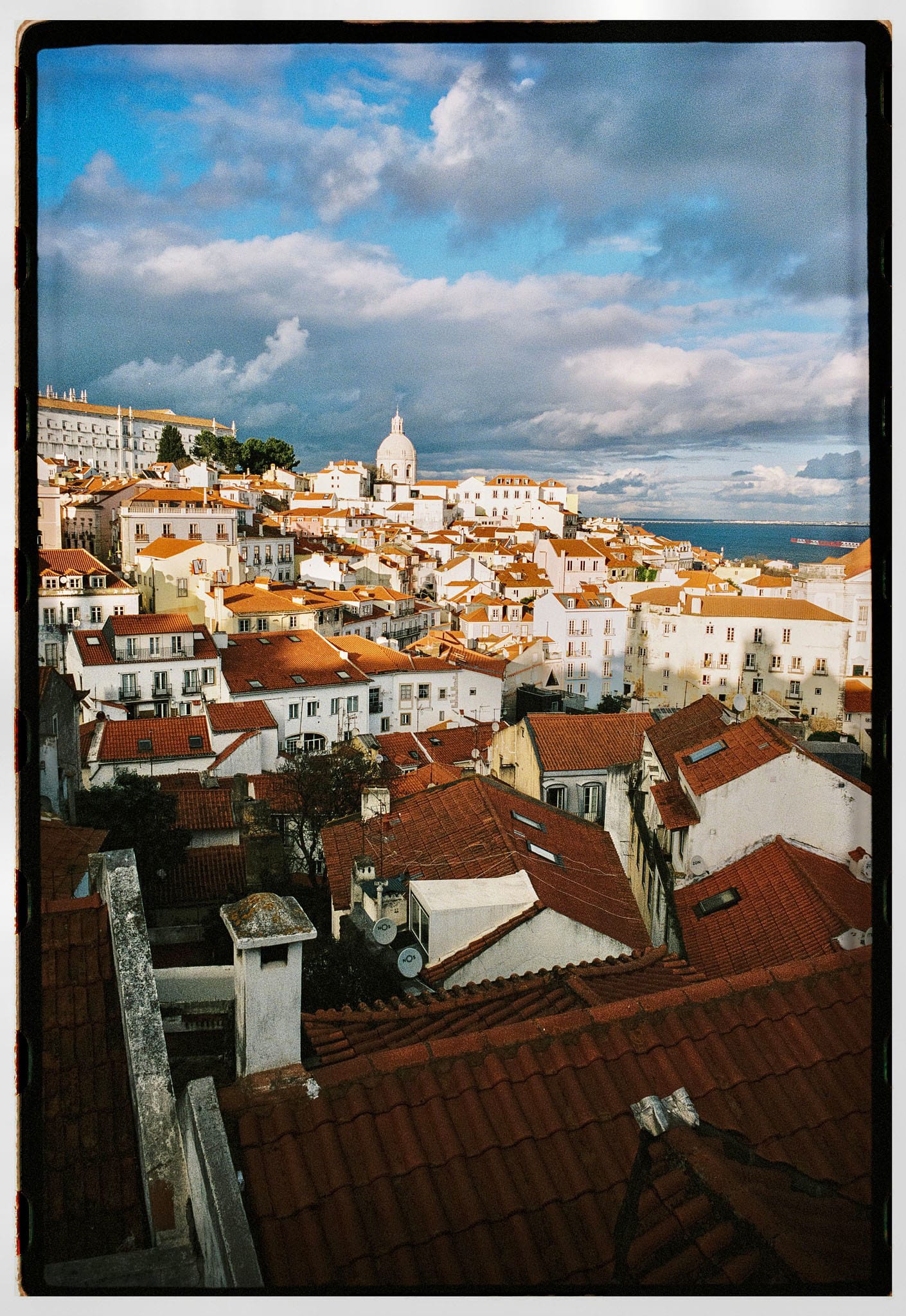 White buildings with terracotta roofs cascading down hillside toward blue water under cloudy sky.