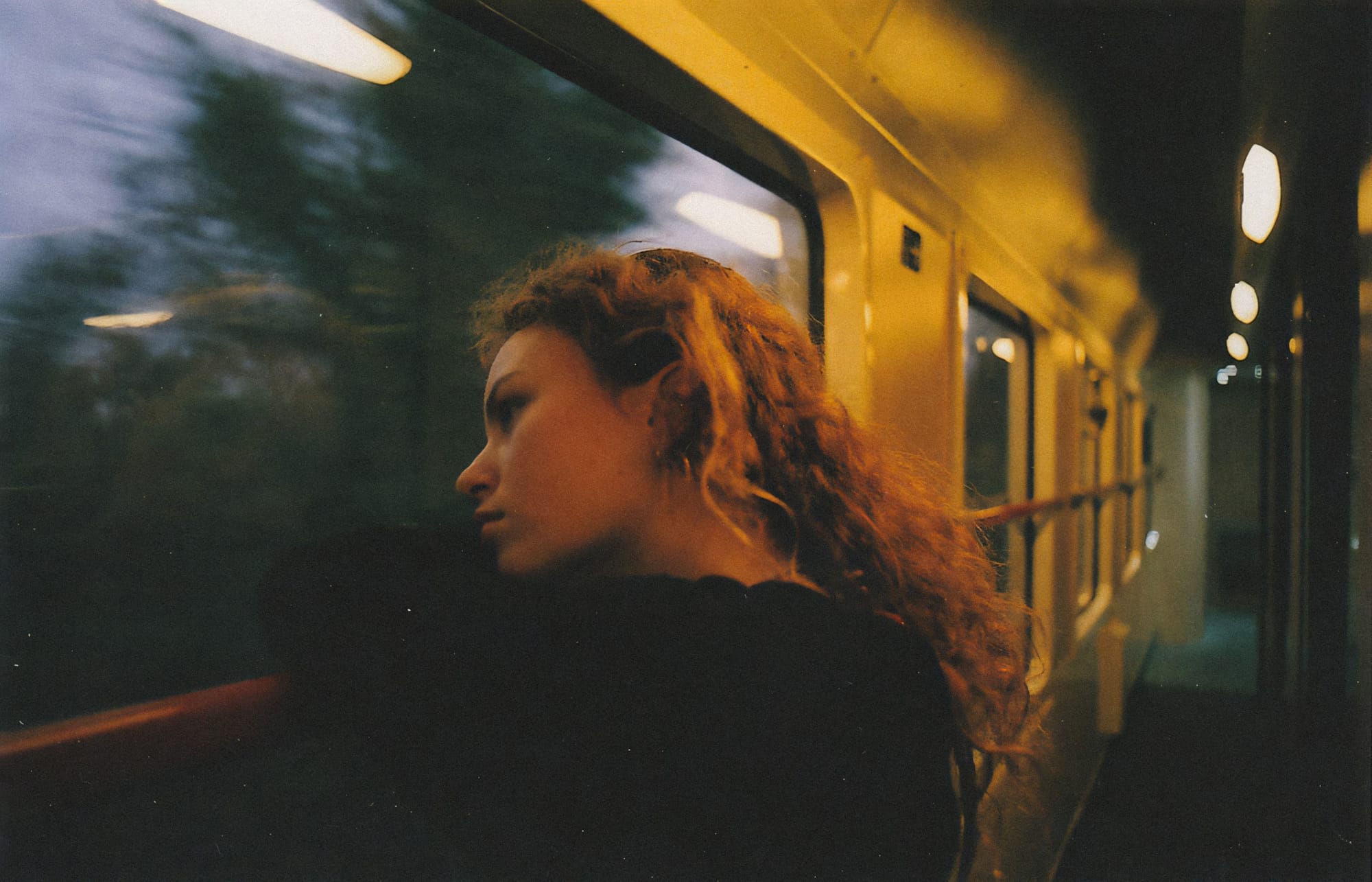 Young woman with curly hair looking out train window at dusk.