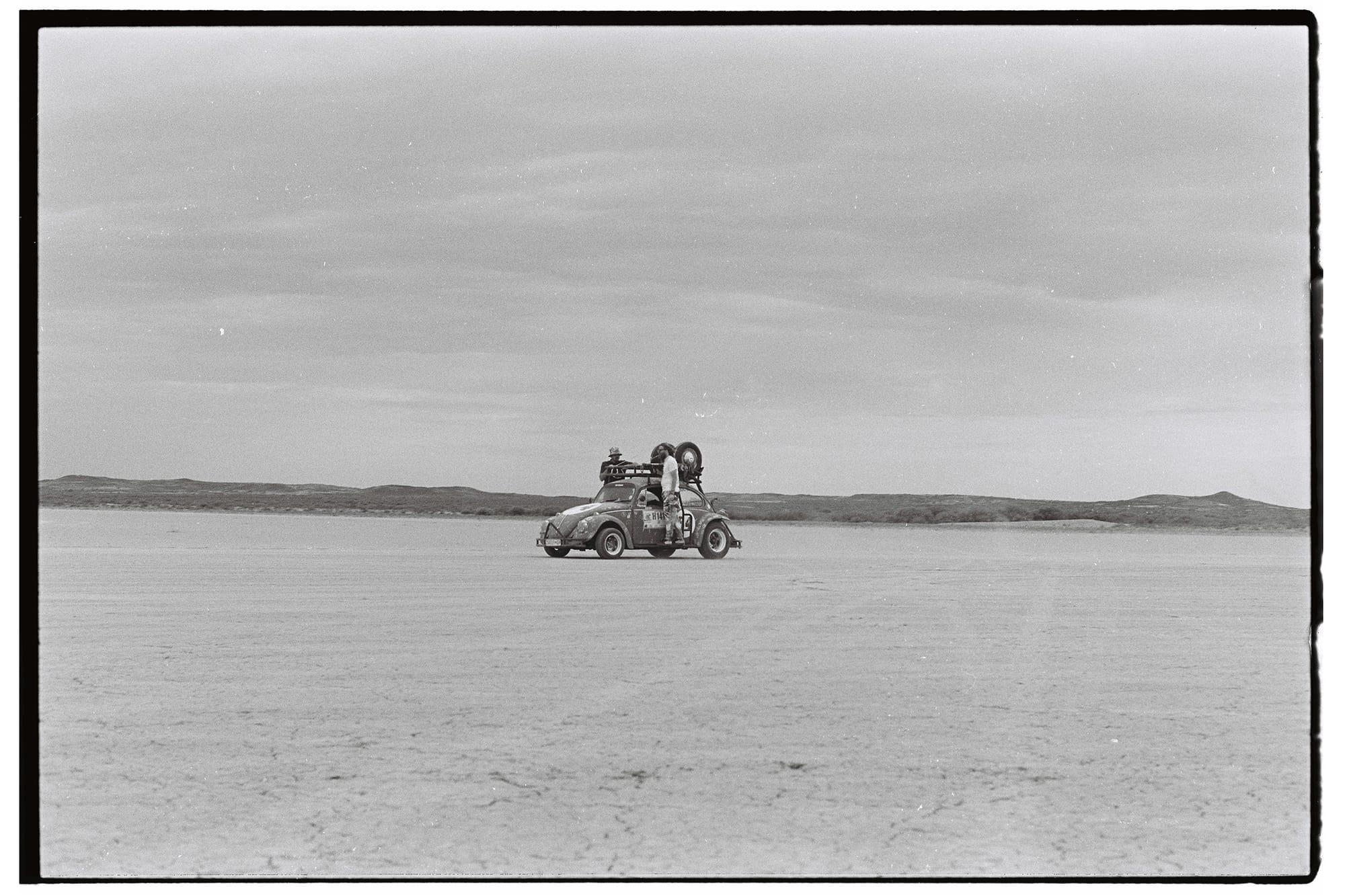 Small sedan with passengers hanging on to its sides crossing vast salt pan.