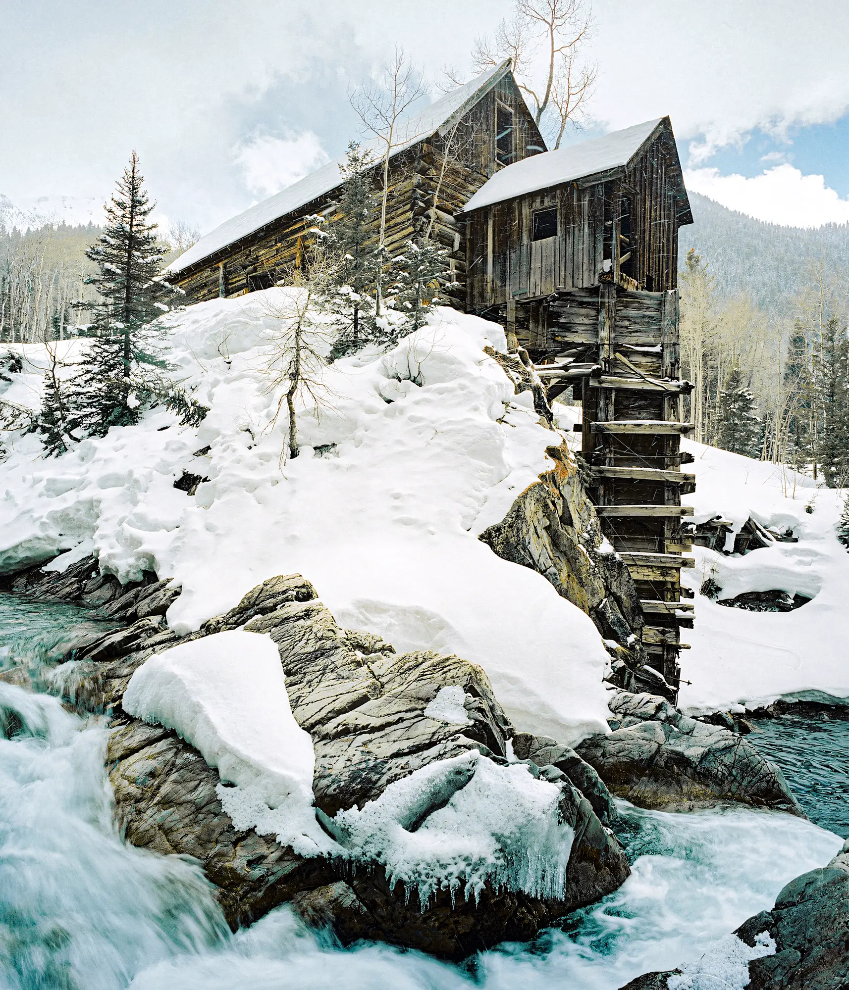 Weathered wooden mill structure beside rushing turquoise stream with deep snow covering rocks and evergreens.