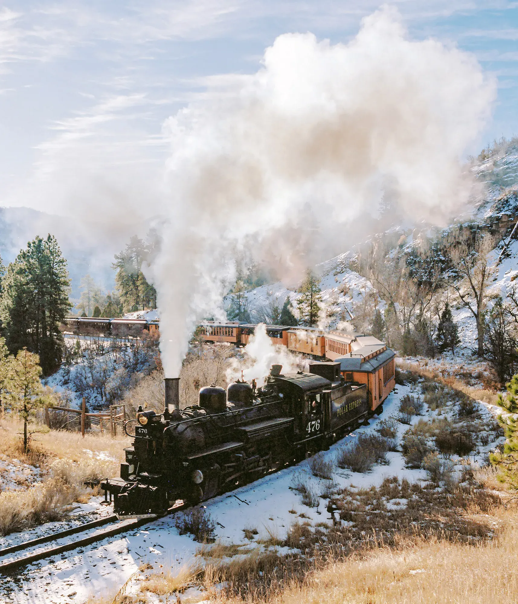 Black steam locomotive crossing mountain canyon on wooden trestle bridge surrounded by snow and evergreens.
