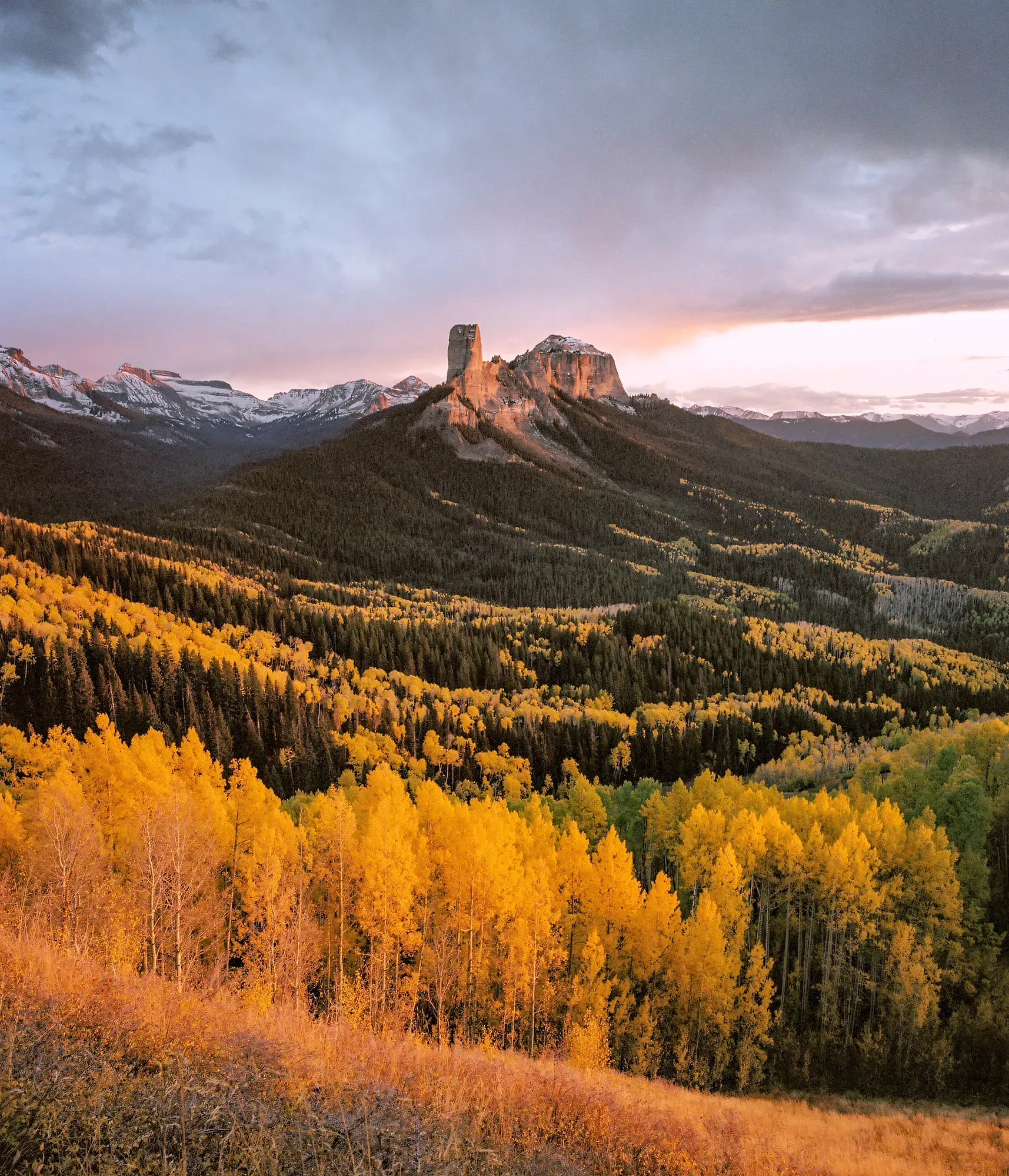 Tall rock formation rising from hillside covered in golden aspens with snow-covered peaks behind.