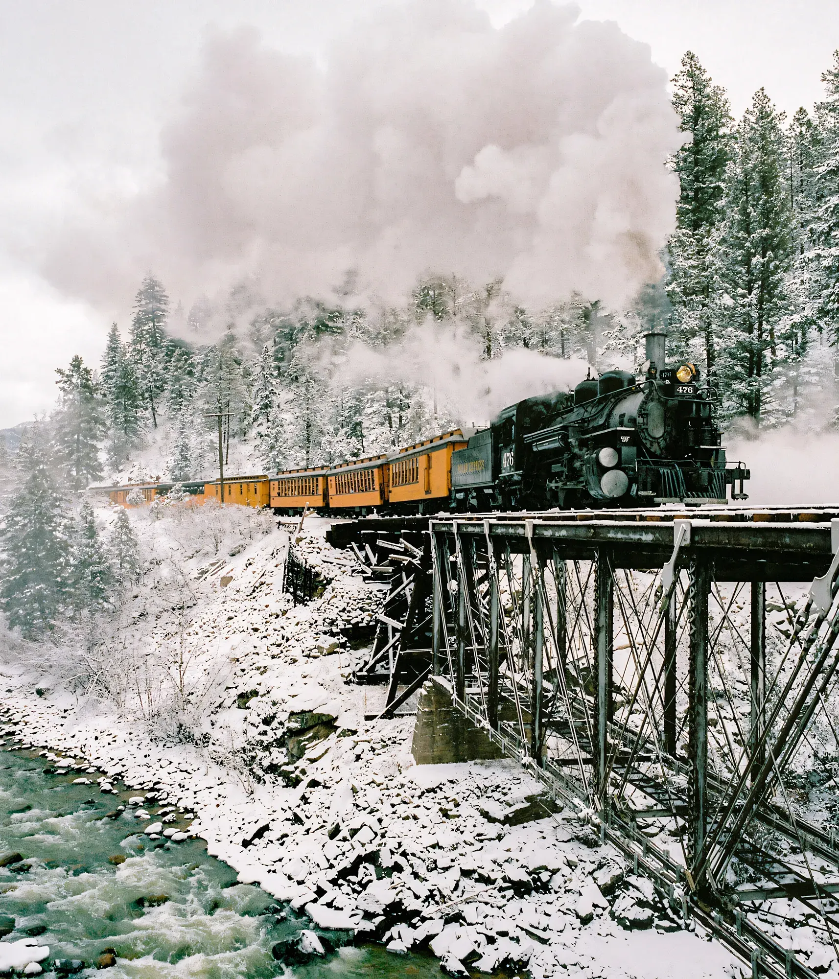 Yellow and black steam train with passenger cars crossing high metal trestle over snowy canyon.