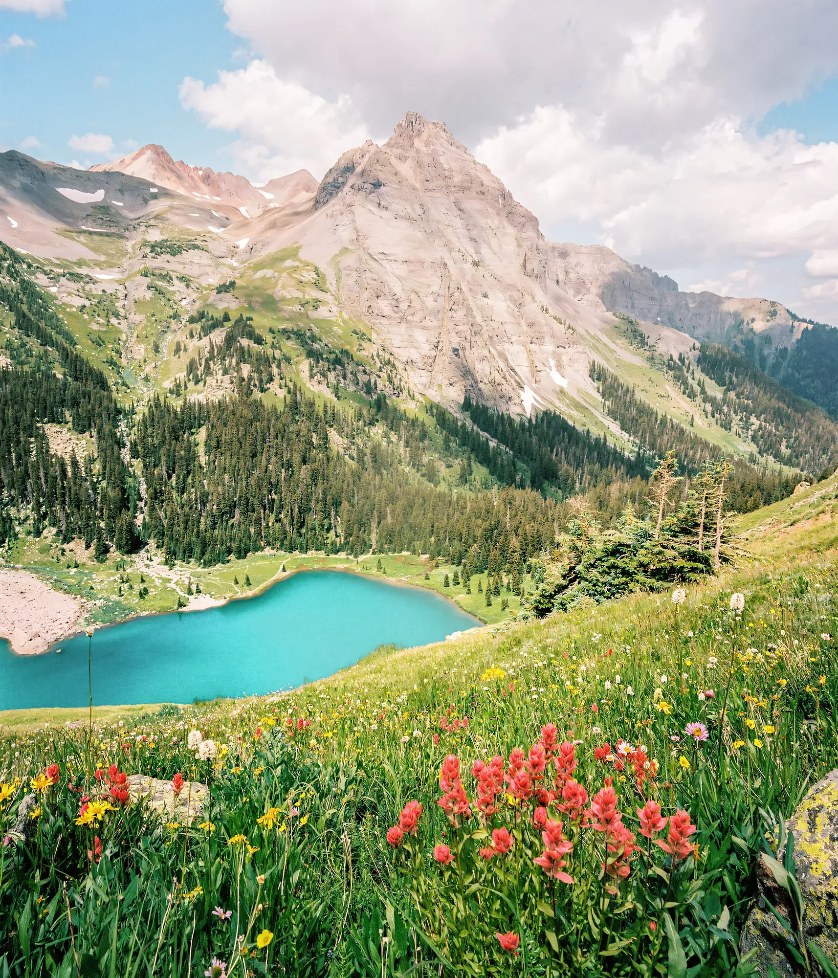 Turquoise alpine lake surrounded by wildflower meadow below rocky mountain peak with remaining snowfields.