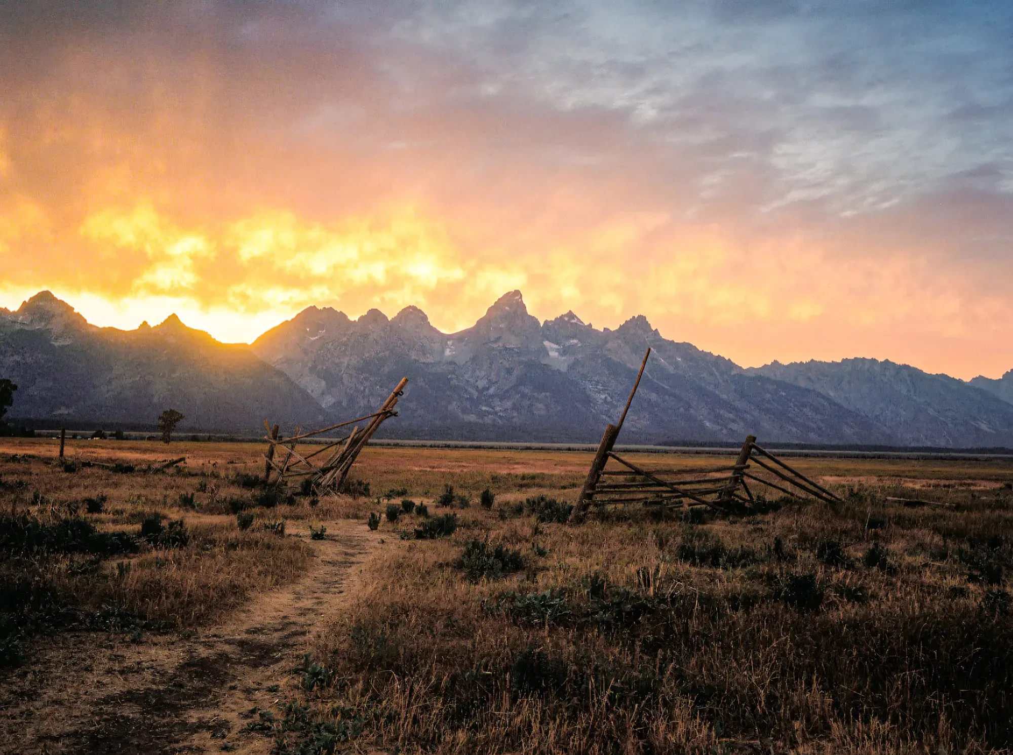 Collapsed wooden fence in prairie at sunrise with Teton mountain range silhouetted against orange sky.
