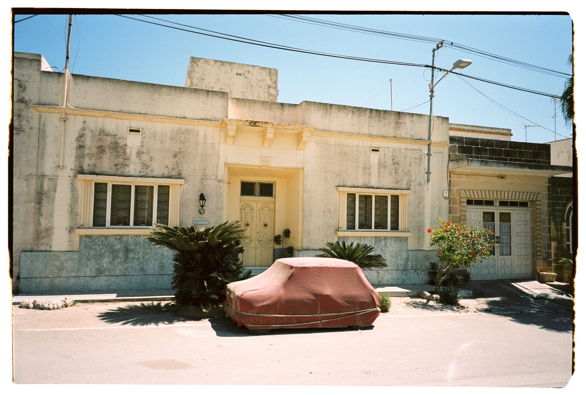 Weathered stucco house with blue-gray lower walls, covered car in courtyard, power lines overhead.