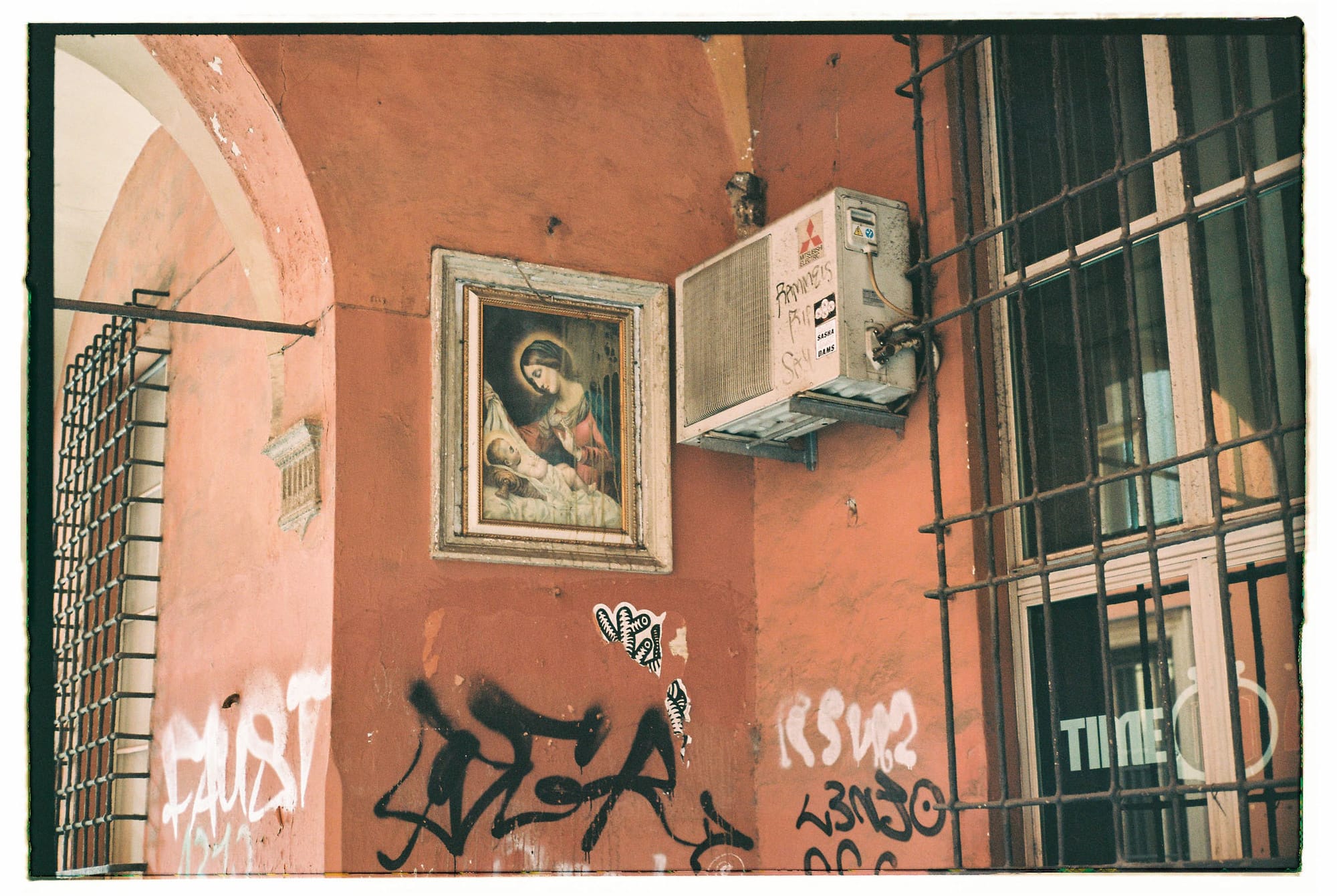 Religious painting of Madonna and child on salmon wall with graffiti tags and mounted utility box.
