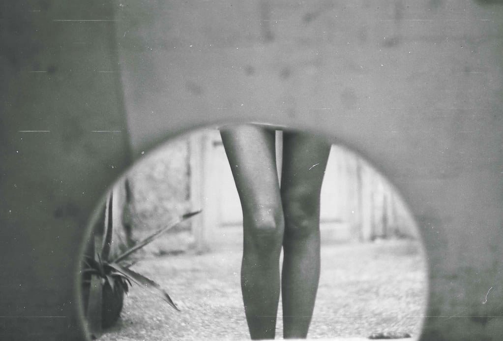 Bare legs of a young woman standing in front of circular mirror, film scratches and dust visible.