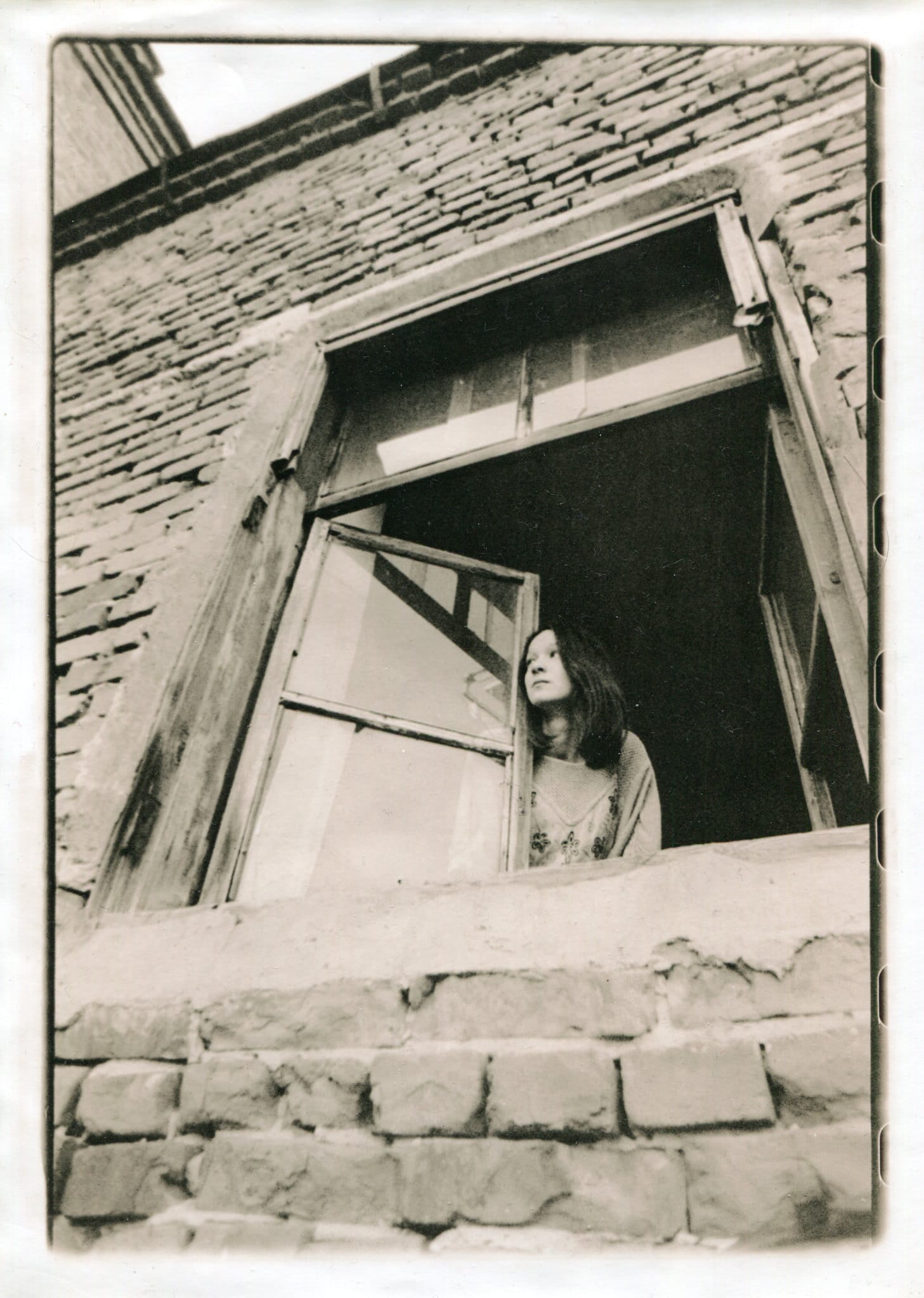 Woman looking upward from open window in brick building photographed from low angle, damaged print edges.
