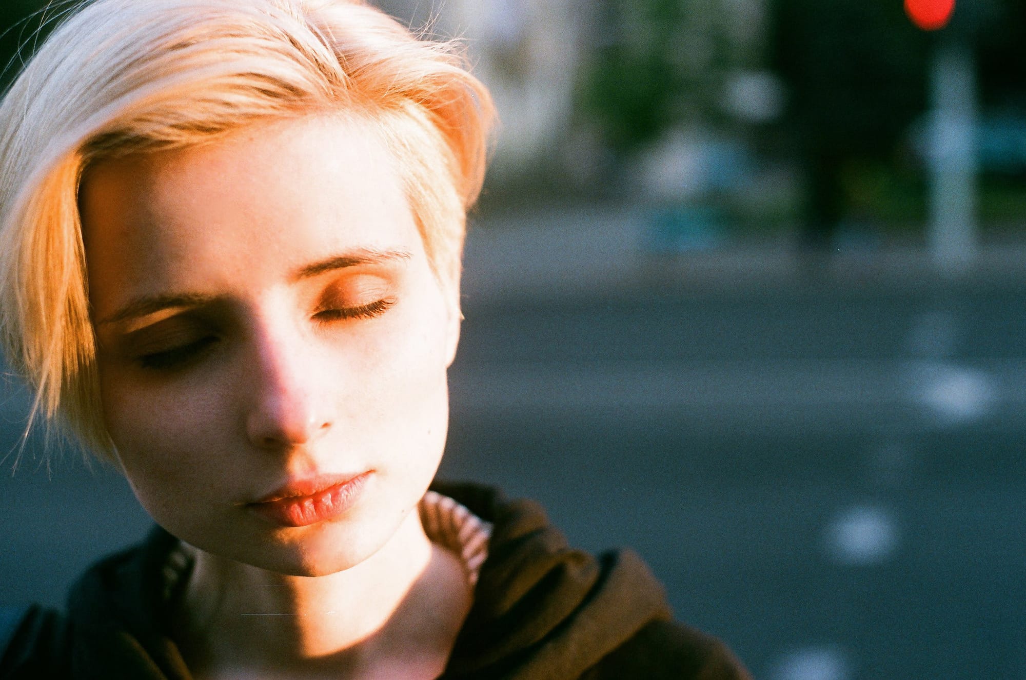 Blonde with closed eyes in warm sunlight against blurred urban background.