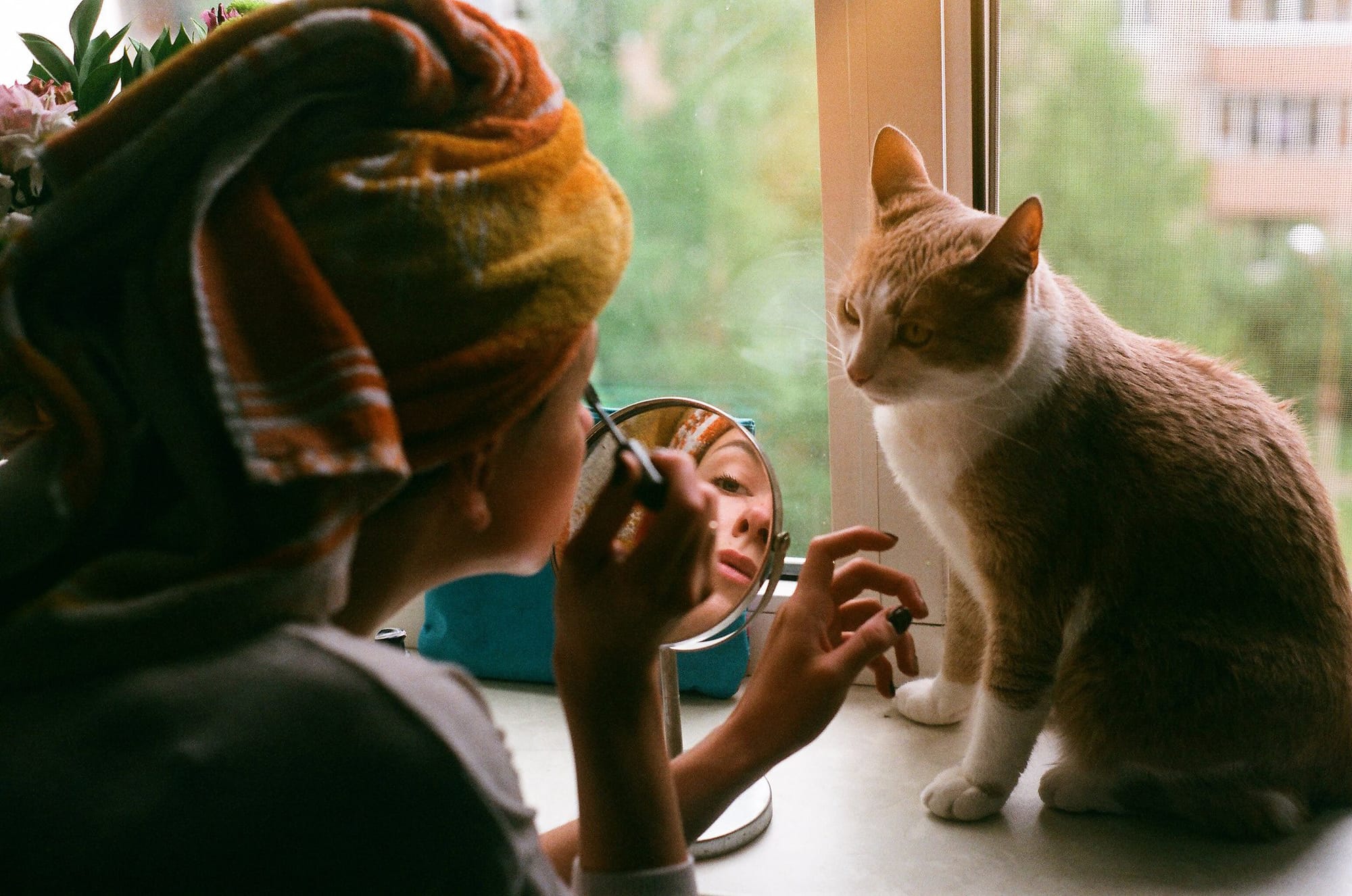 Girl applying makeup with compact mirror at window sill, orange tabby cat watching nearby.