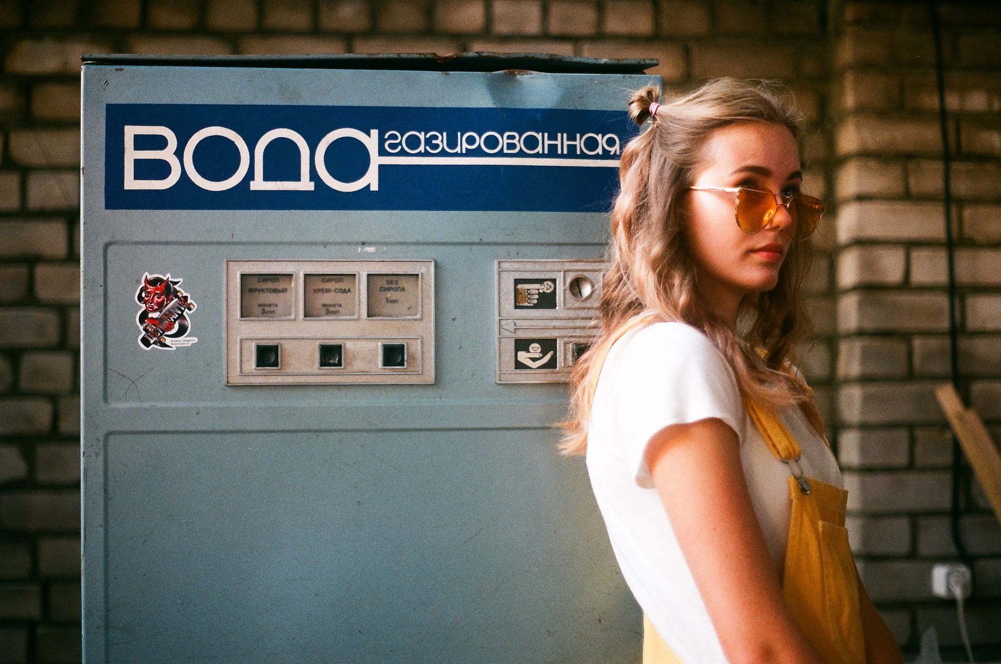Blonde in orange sunglasses and yellow overalls standing beside Soviet-era infrastructure with Cyrillic text.