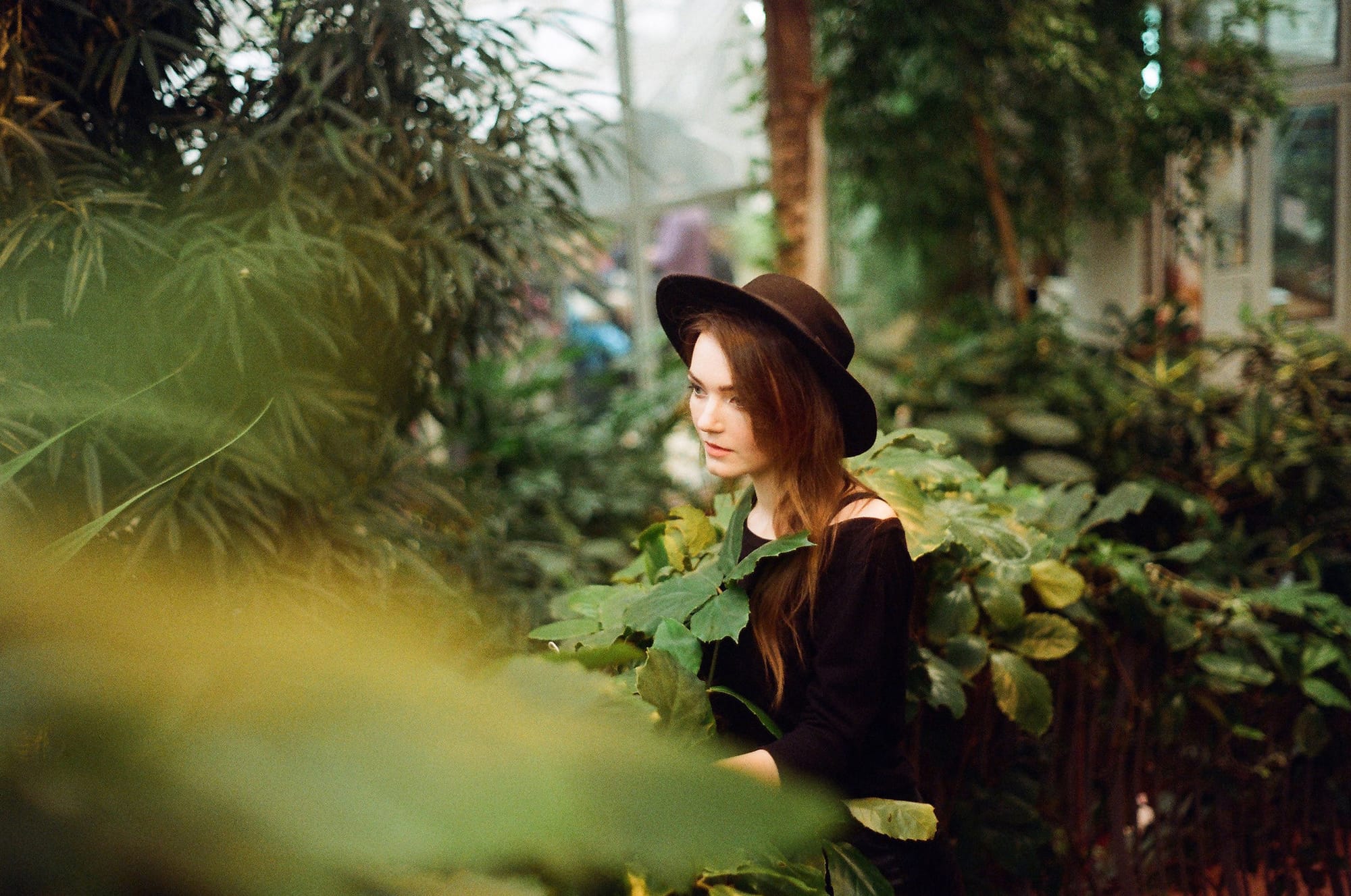 Brunette in black hat and off-shoulder top surrounded by dense greenhouse plants and foliage.