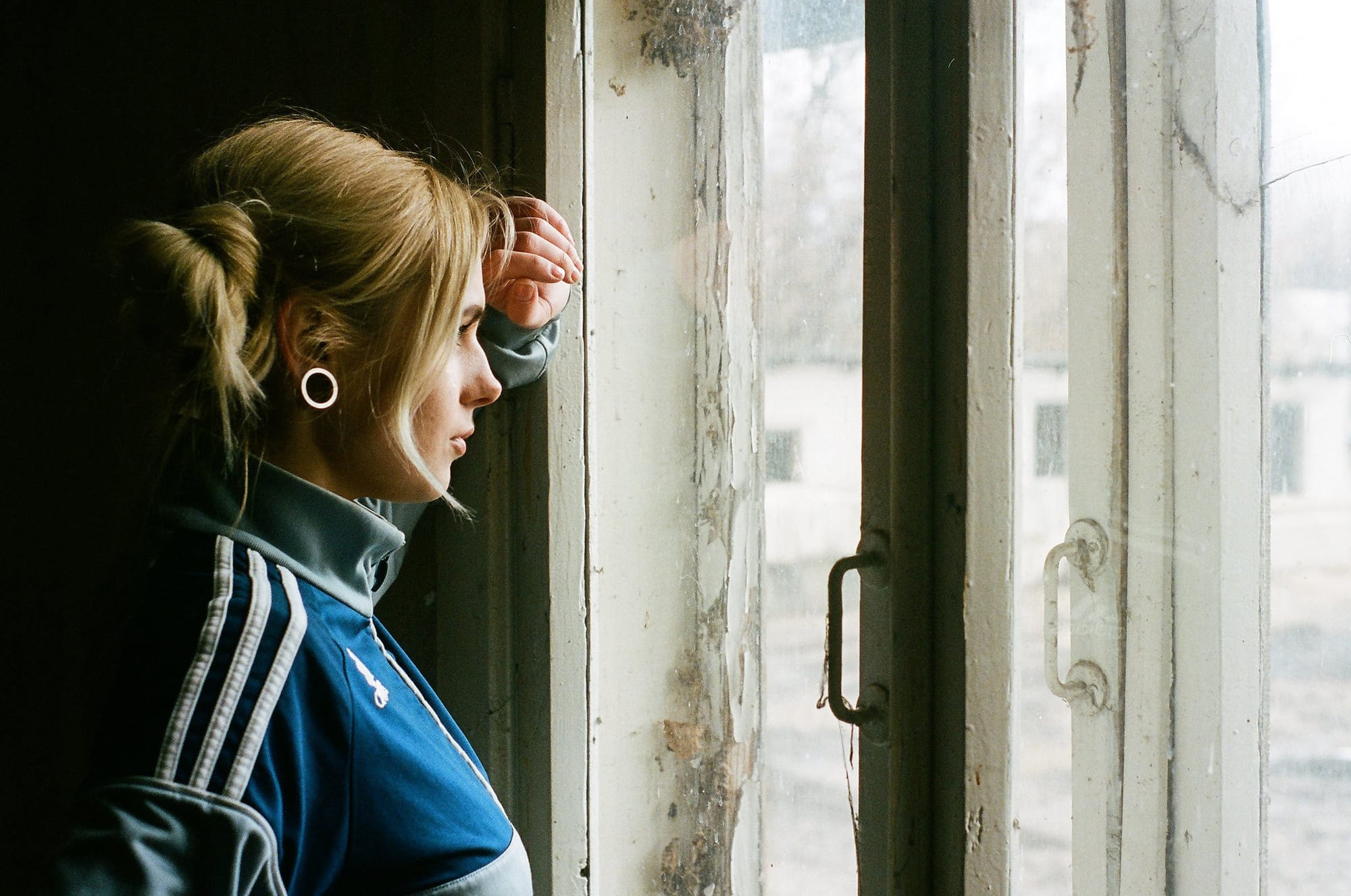 Blonde in blue Adidas track jacket leaning against weathered window frame with circular earring.