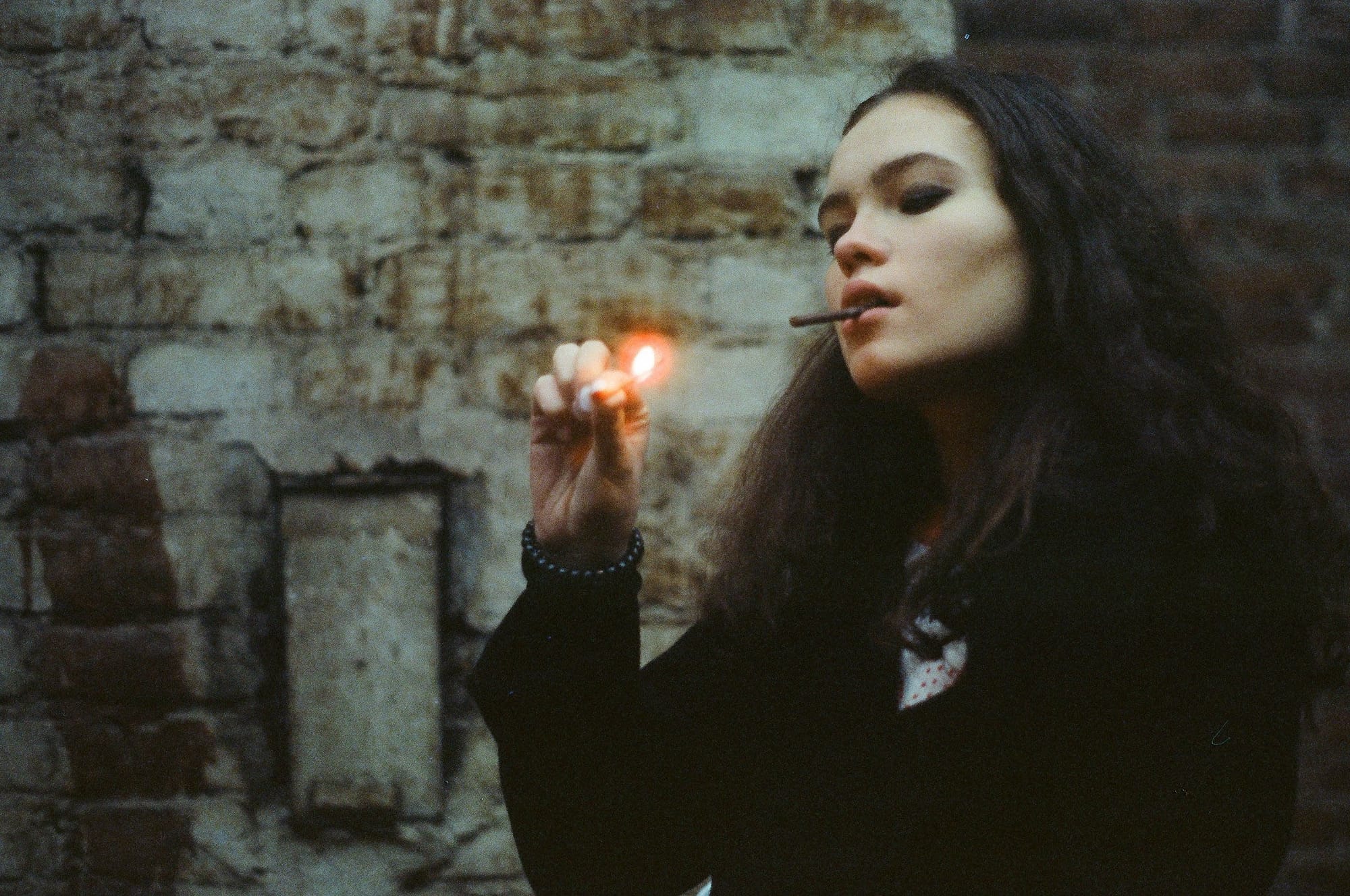 Woman lighting cigarette with flame visible near face against deteriorating brick wall.