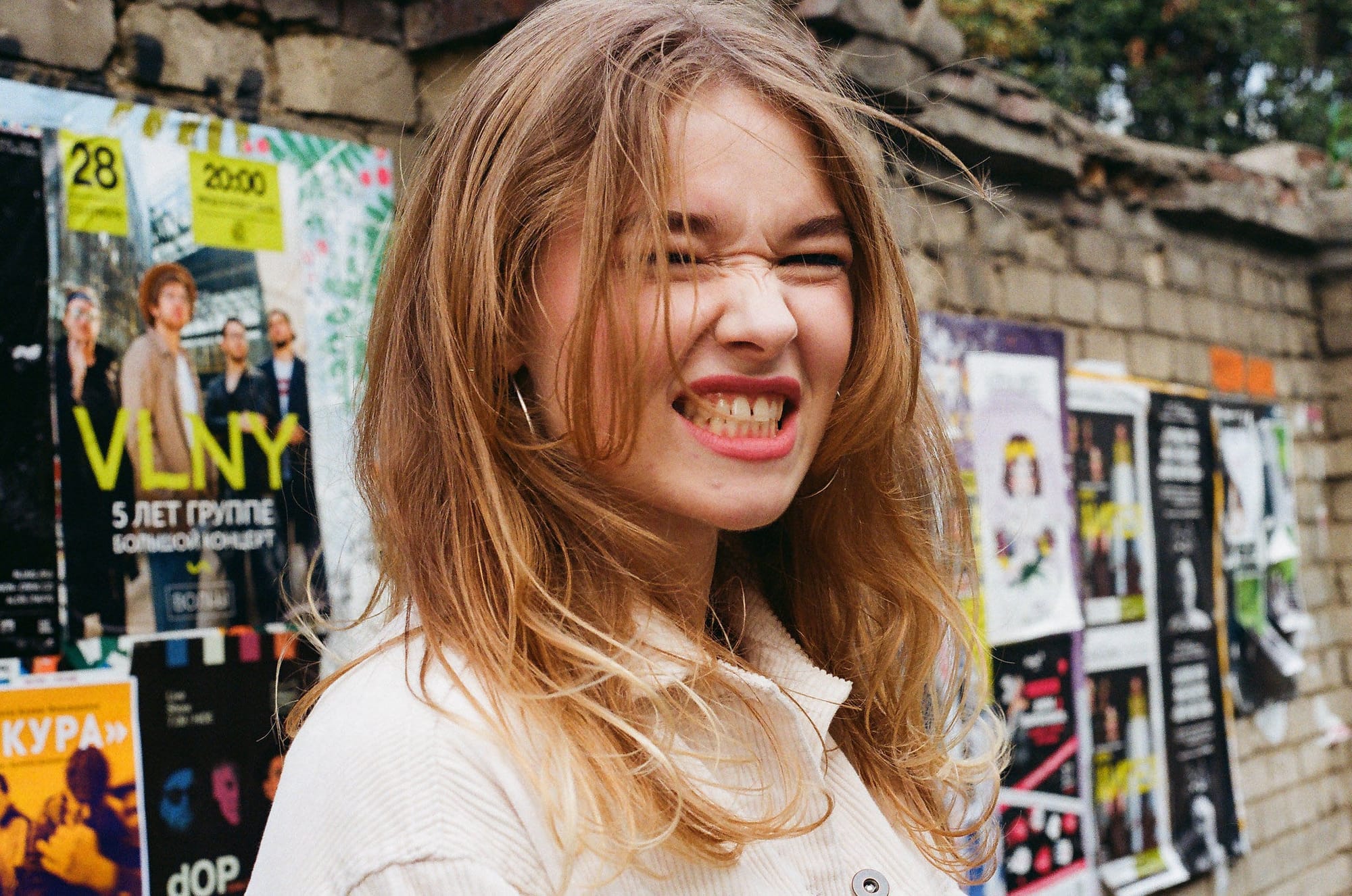 Girl making faces with windblown hair in front of poster-covered brick wall with Cyrillic text.