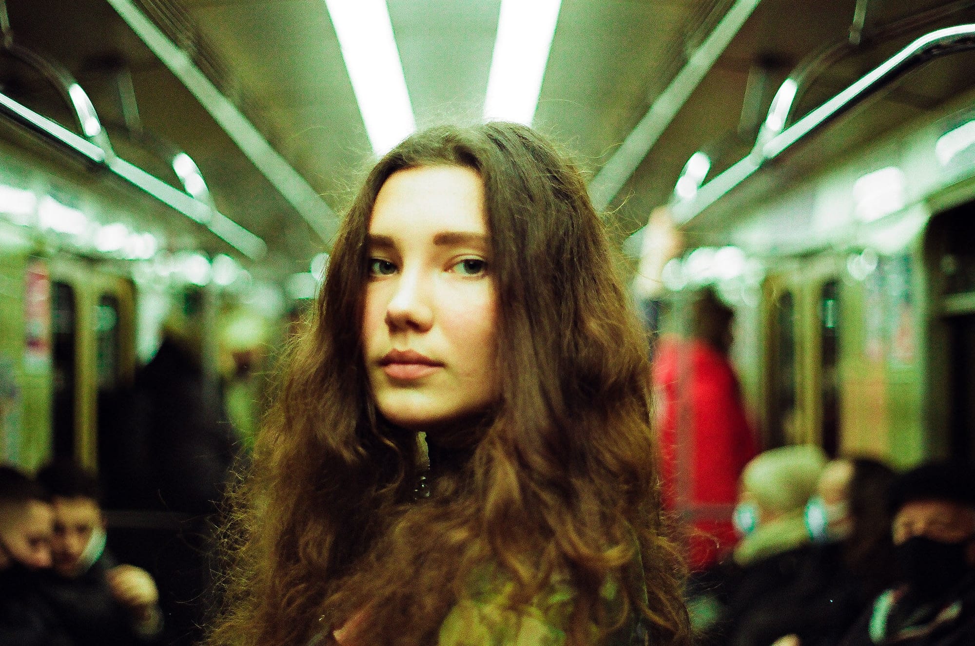 Girl with long brown hair in subway car with green fluorescent ceiling lights.