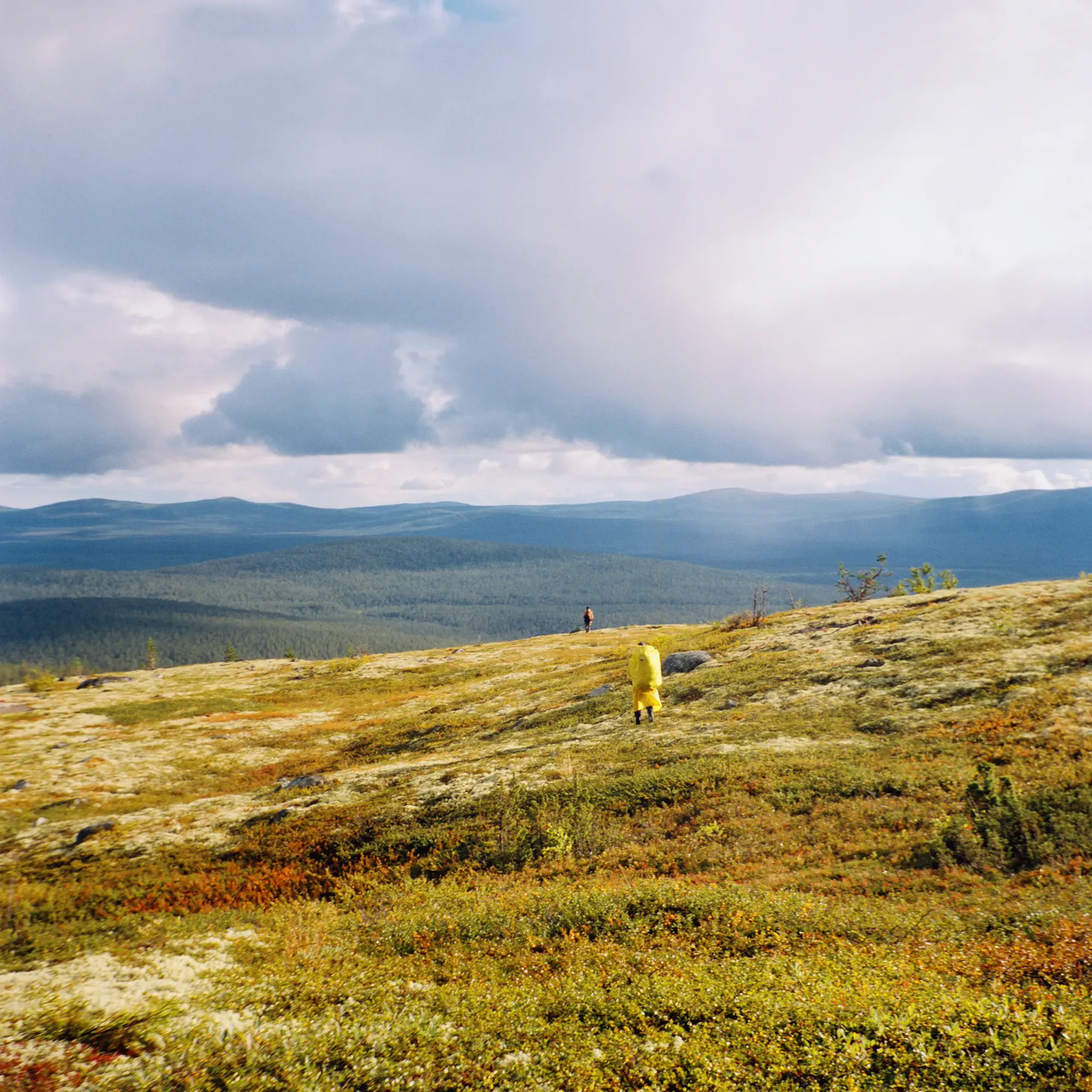 Two people walking across golden tundra hillside beneath stormy mountain sky.