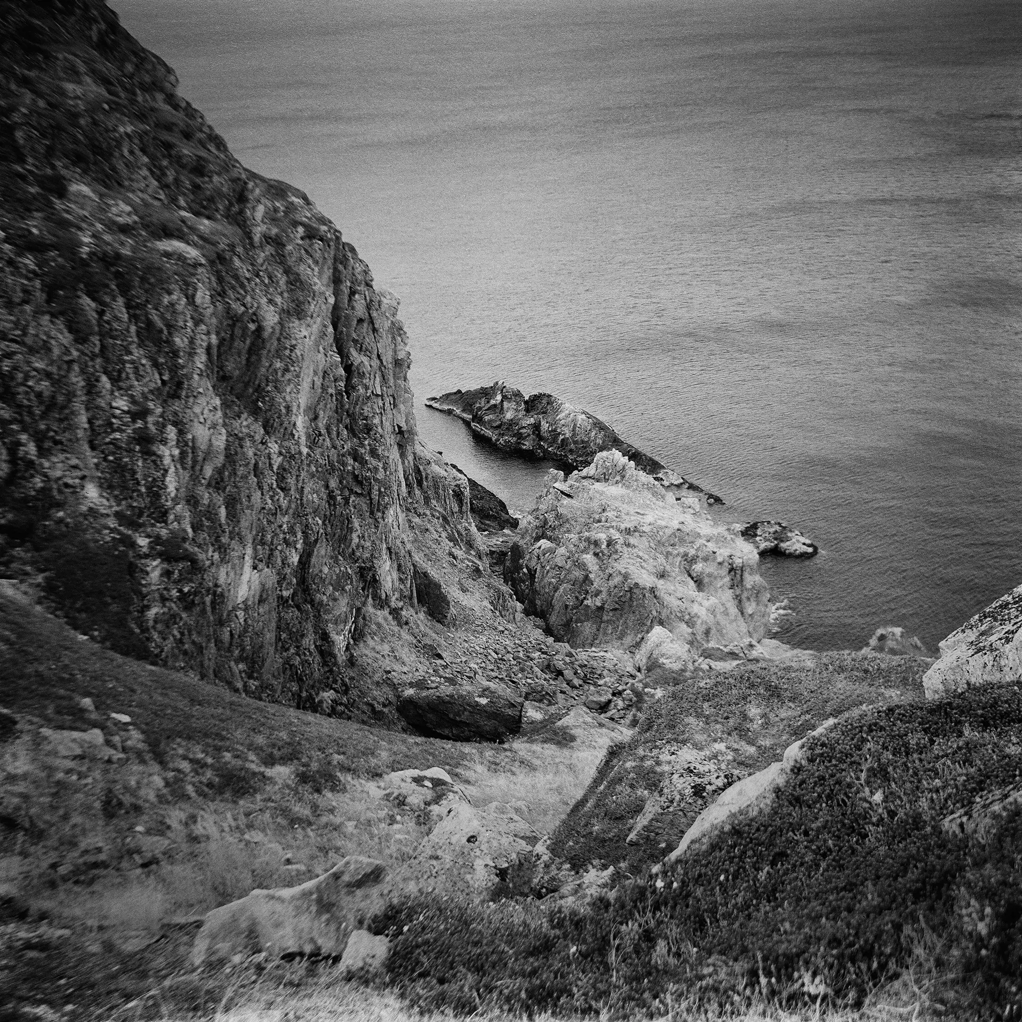 Black and white coastal cliff face with eroded rock formations jutting into calm sea.