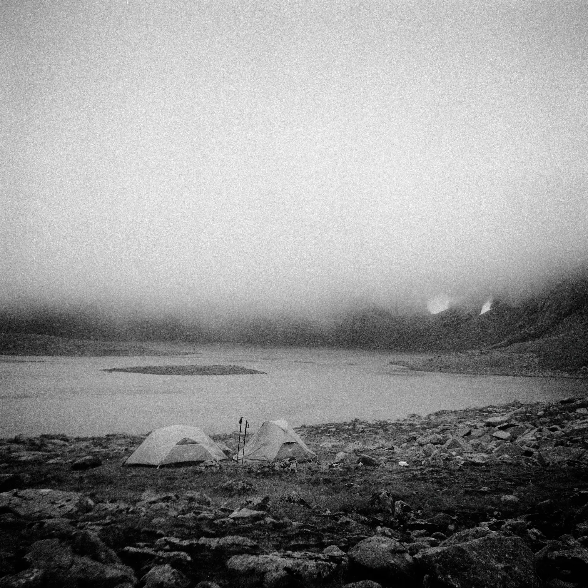Two camping tents on rocky shore beside lake with fog-obscured mountain in background.