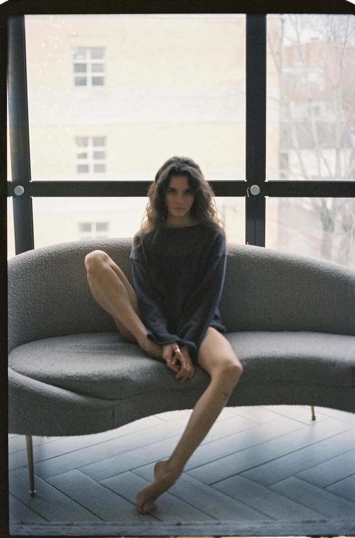 Woman in oversized sweater sits on curved gray couch by window overlooking buildings.