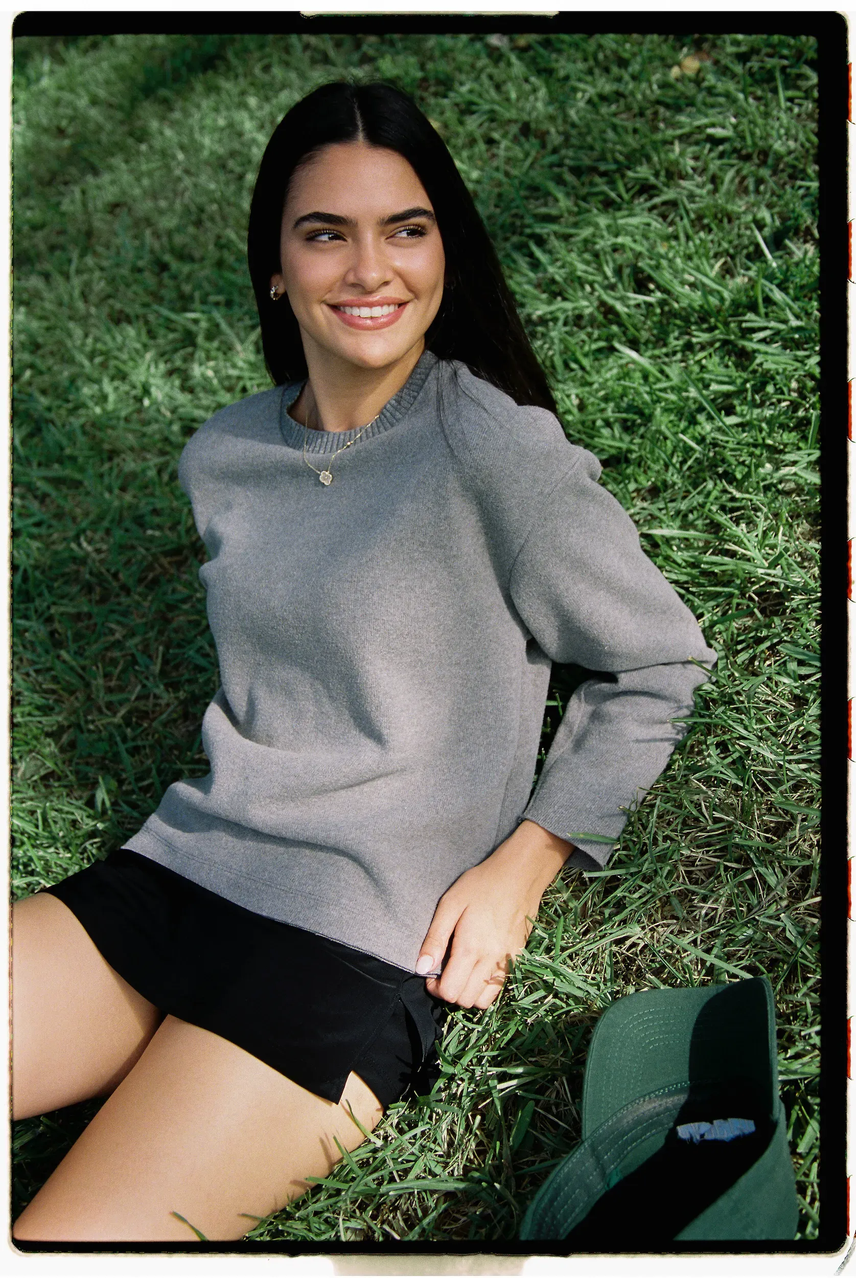 Brunette in gray sweater and heart necklace smiling while sitting on grass in bright sunlight.