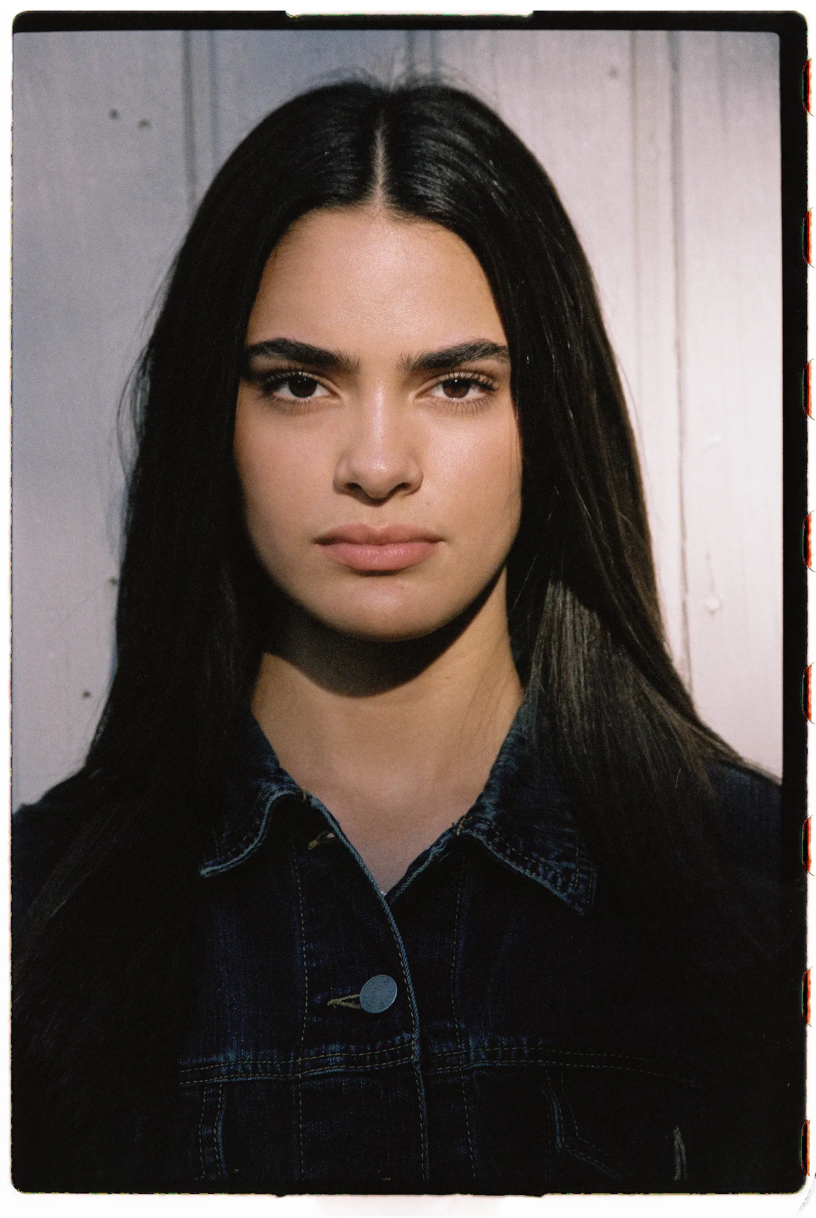 Frontal portrait of a fashion model in dark denim jacket against weathered white painted wall.