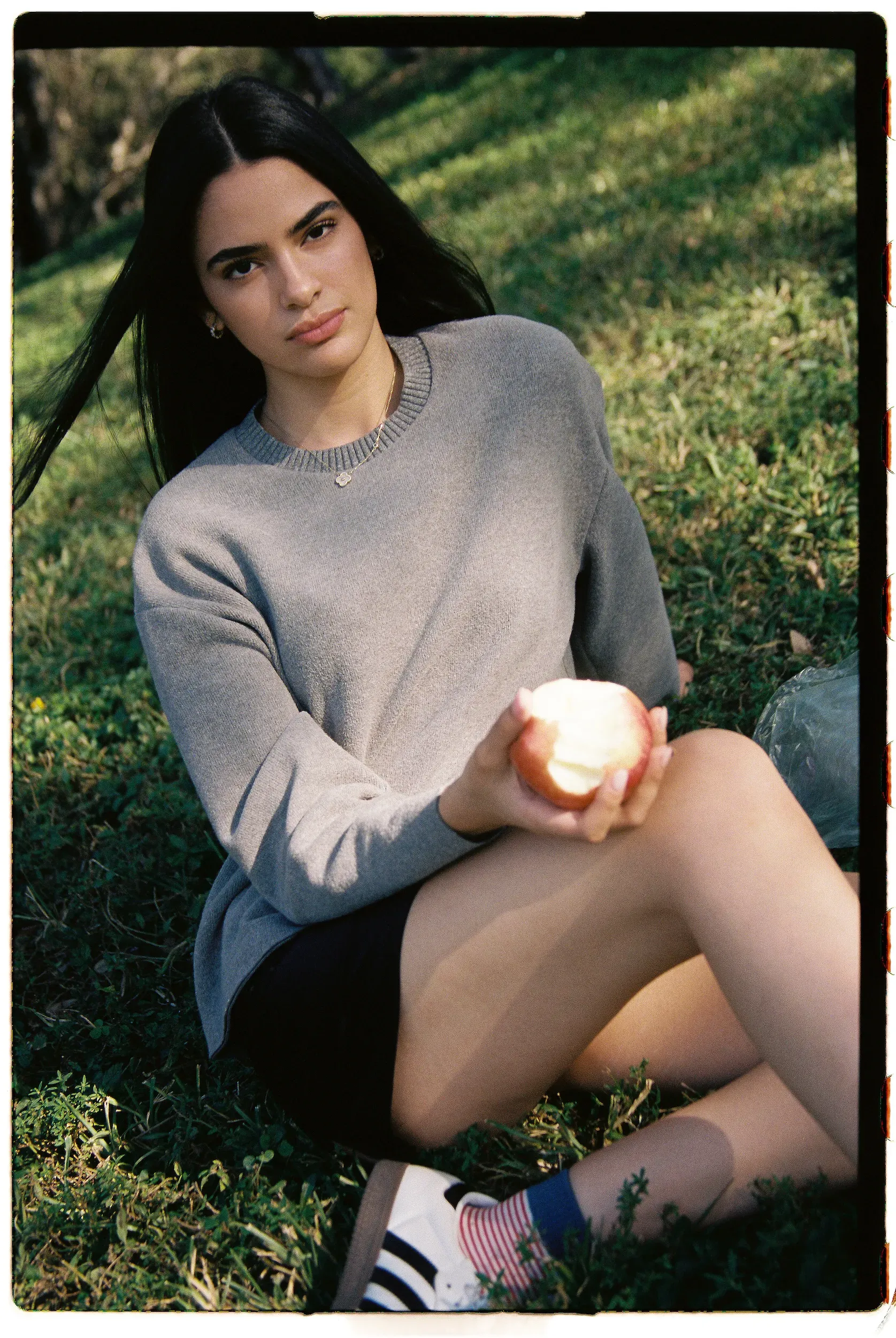 Fashion model in gray sweater holding partially eaten apple while seated on grass with green cap nearby.