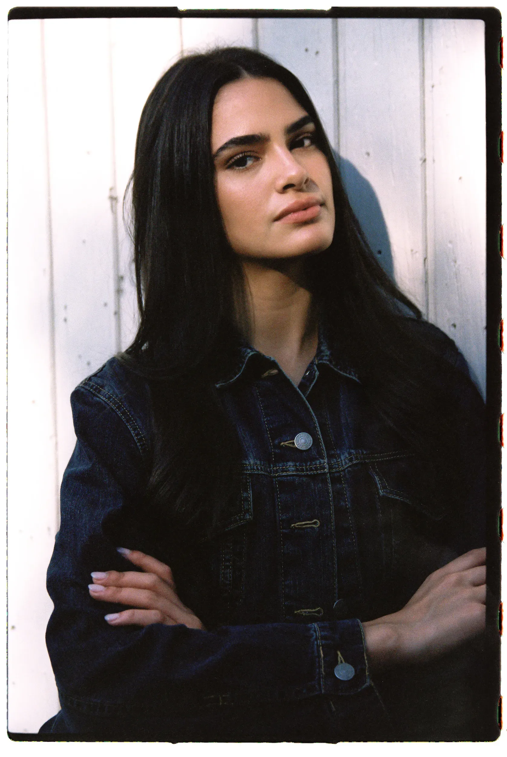 Young woman in dark denim jacket with arms crossed looking upward against white wooden siding.