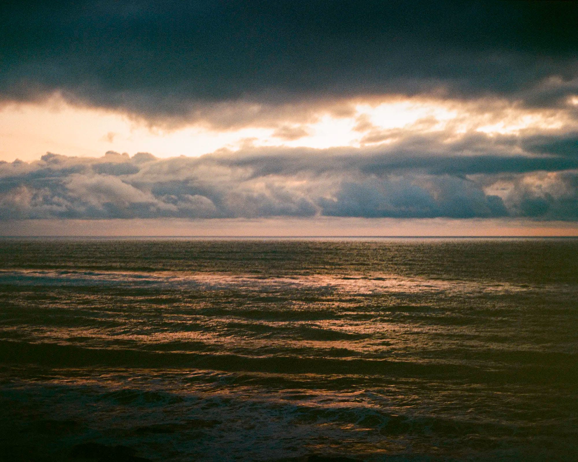 Dark ocean waves at sunset with dramatic storm clouds breaking golden light across water surface.
