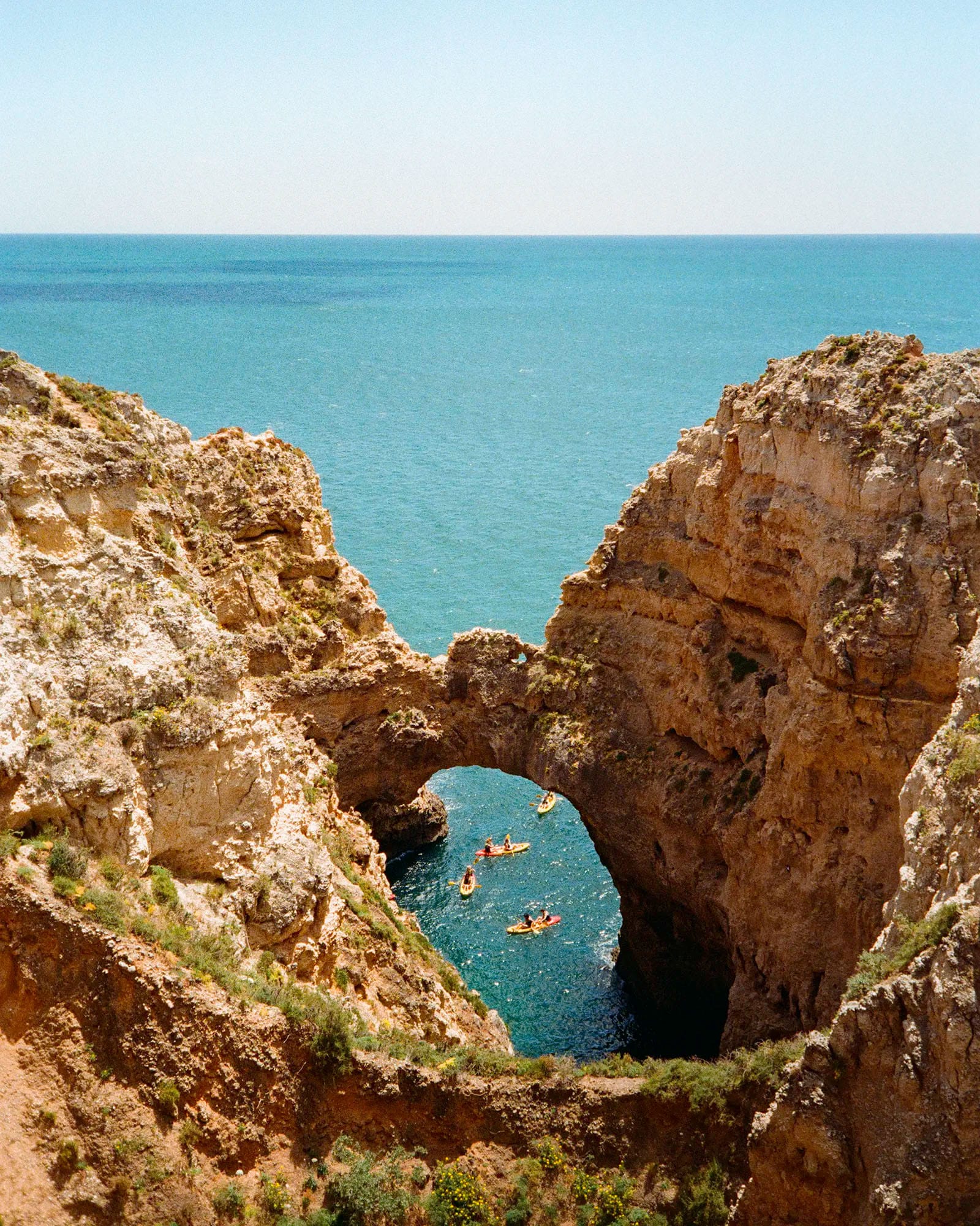 Natural rock arch over turquoise ocean cove with kayakers paddling through opening below weathered cliffs.