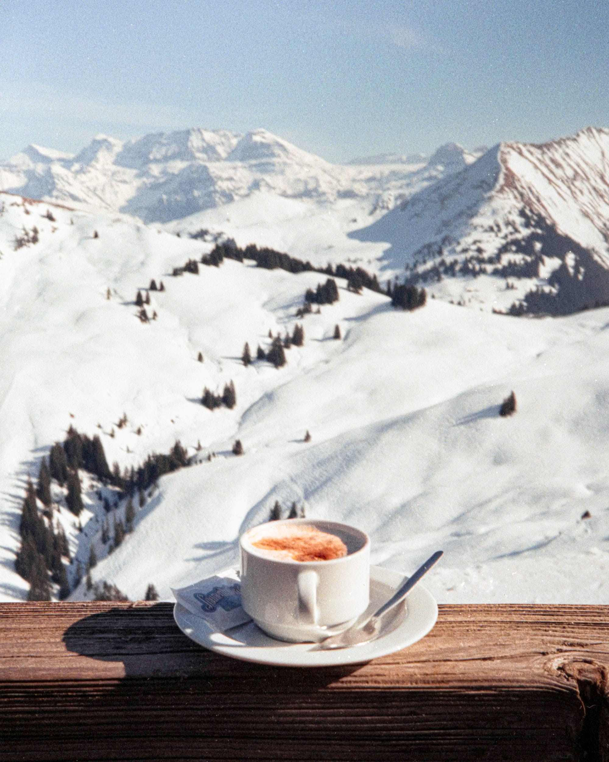 Coffee cup on wooden deck railing overlooking snow-covered Alpine ski slopes and mountain peaks.