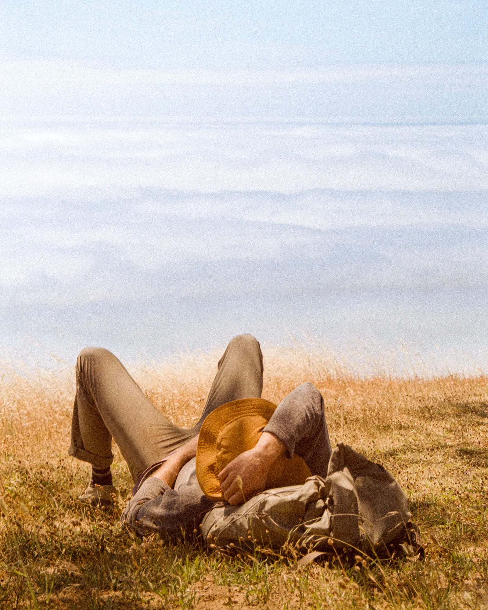 Man lying in grass wearing tan hat overlooking a thick cloud layer.