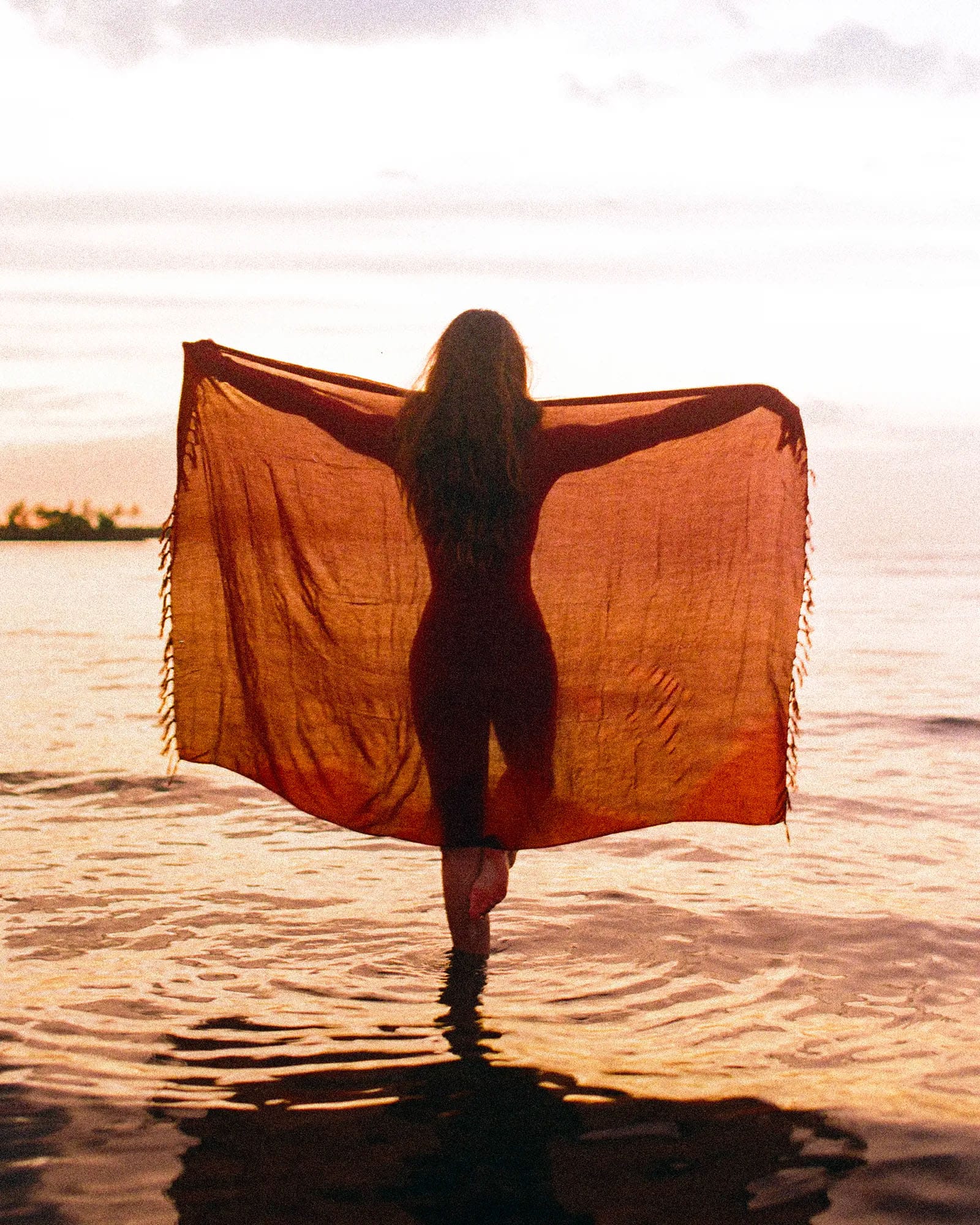 Silhouetted woman holding orange shawl at water's edge during golden hour sunset.