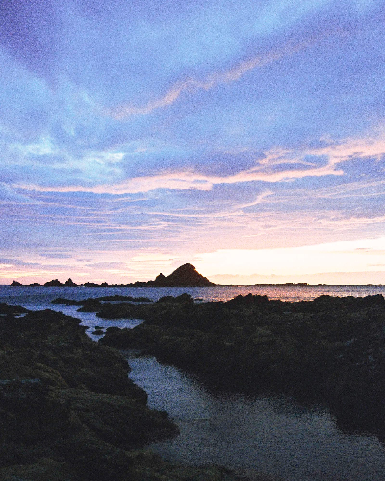 Rocky tidal pools at sunset with island silhouette on horizon and pink-purple clouds.