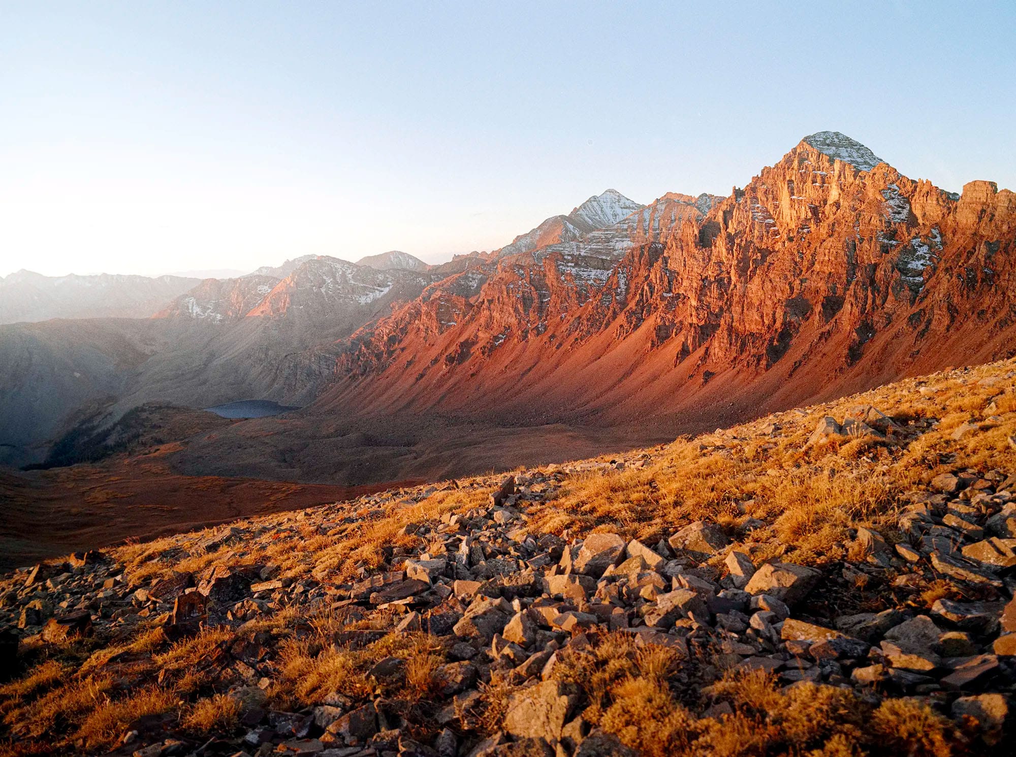 Jagged alpine ridge bathed in sunrise light with small lake and golden scree slope in foreground.