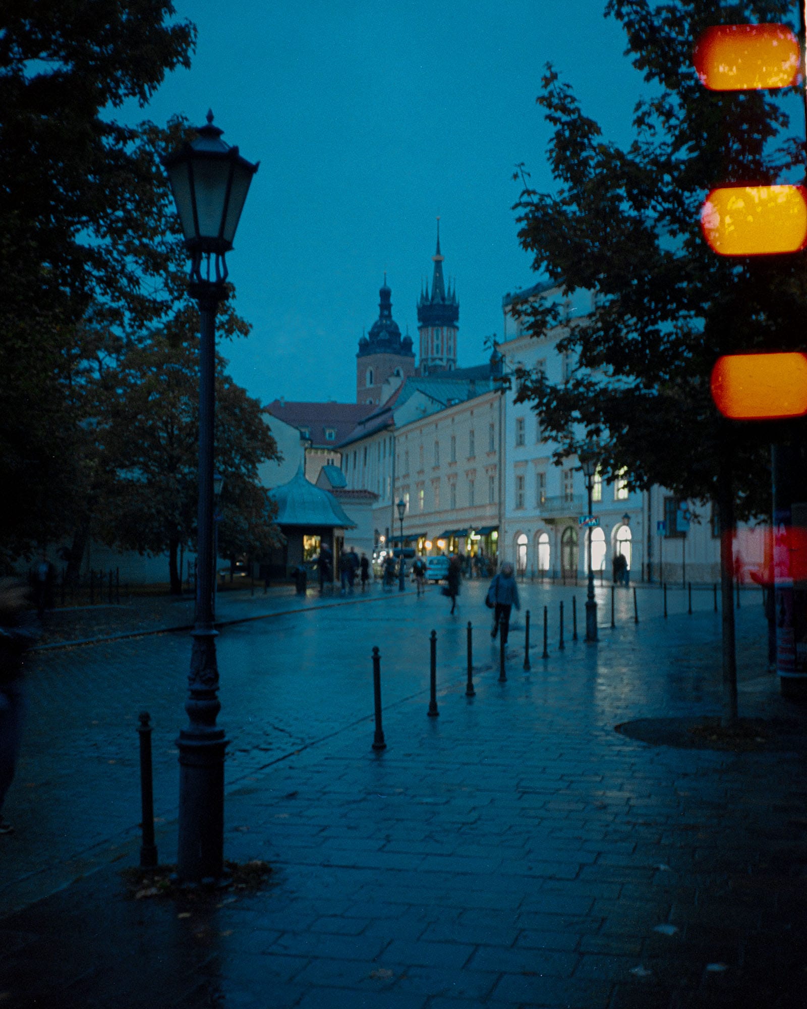 Wet cobblestone plaza at blue hour with illuminated street lamps, historic church towers, and blurred pedestrians under trees.
